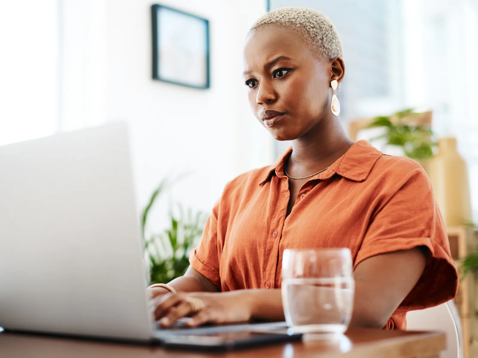 Focused young woman working on laptop at home with a glass of water on the table in a bright room