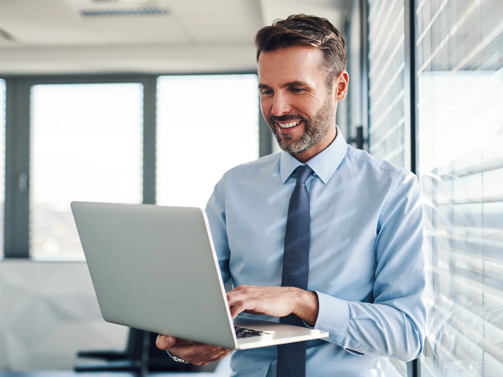 Smiling businessman in blue shirt and tie working on laptop in modern office with natural light.
