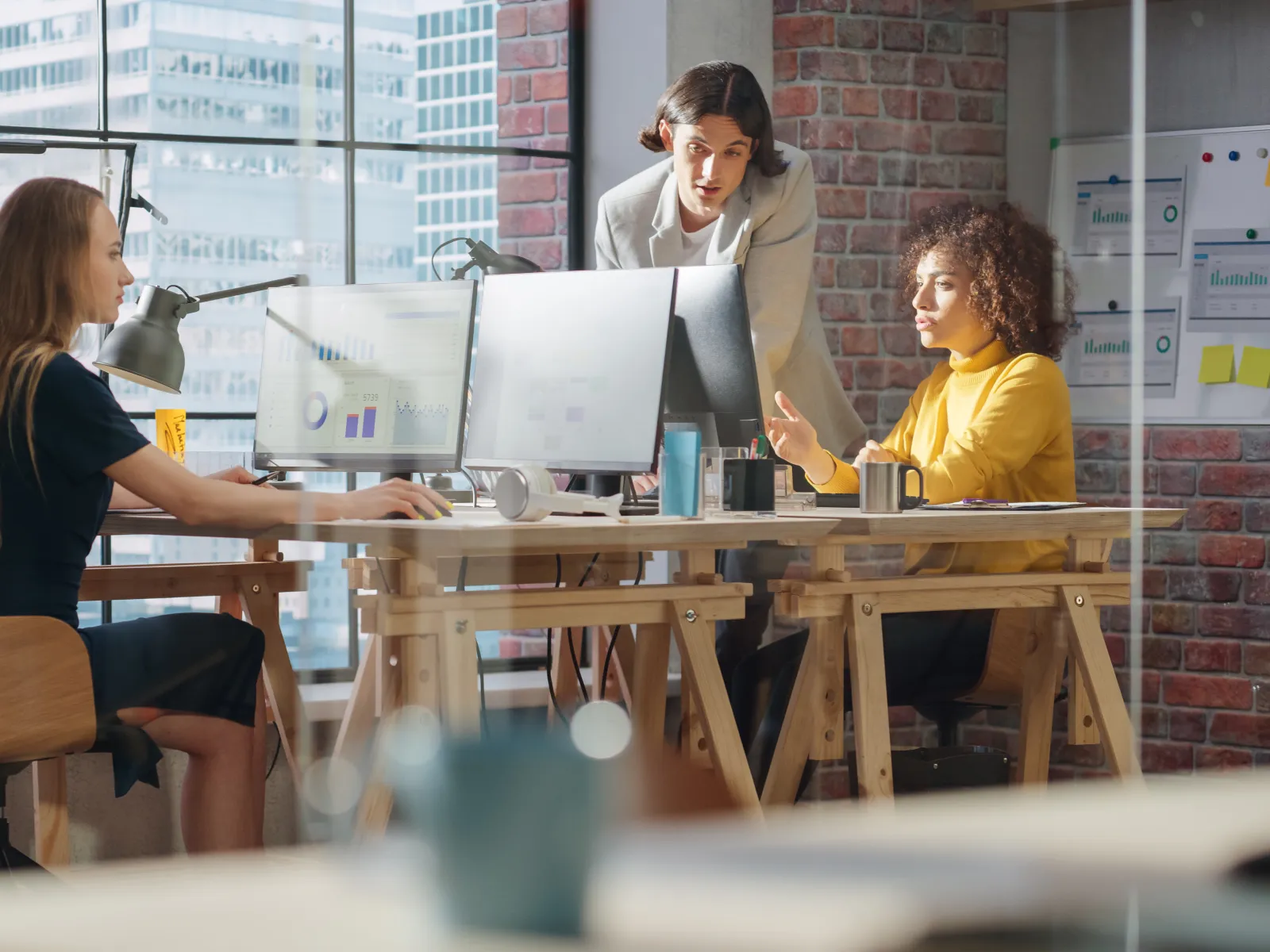 Three women collaborating in a modern office space with computers and city skyline views through large windows