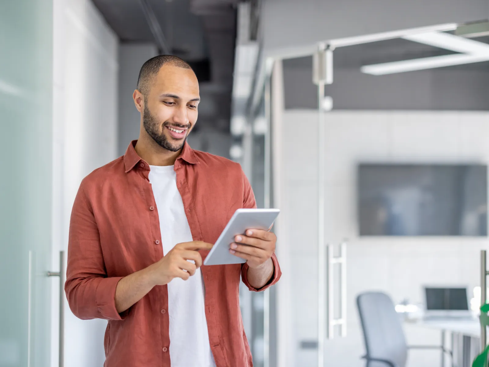 Smiling man using digital tablet in modern office corridor with glass walls and conference room.
