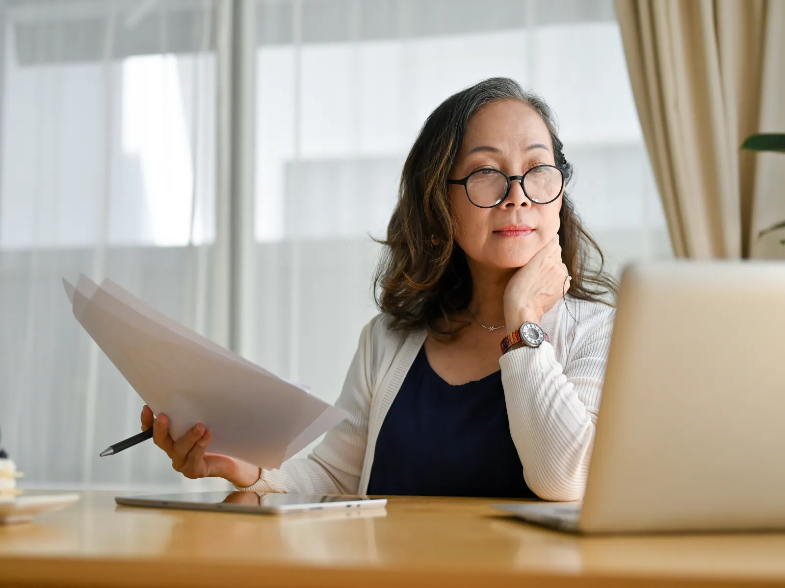 Mature woman wearing glasses reviewing documents while working on laptop at home office desk