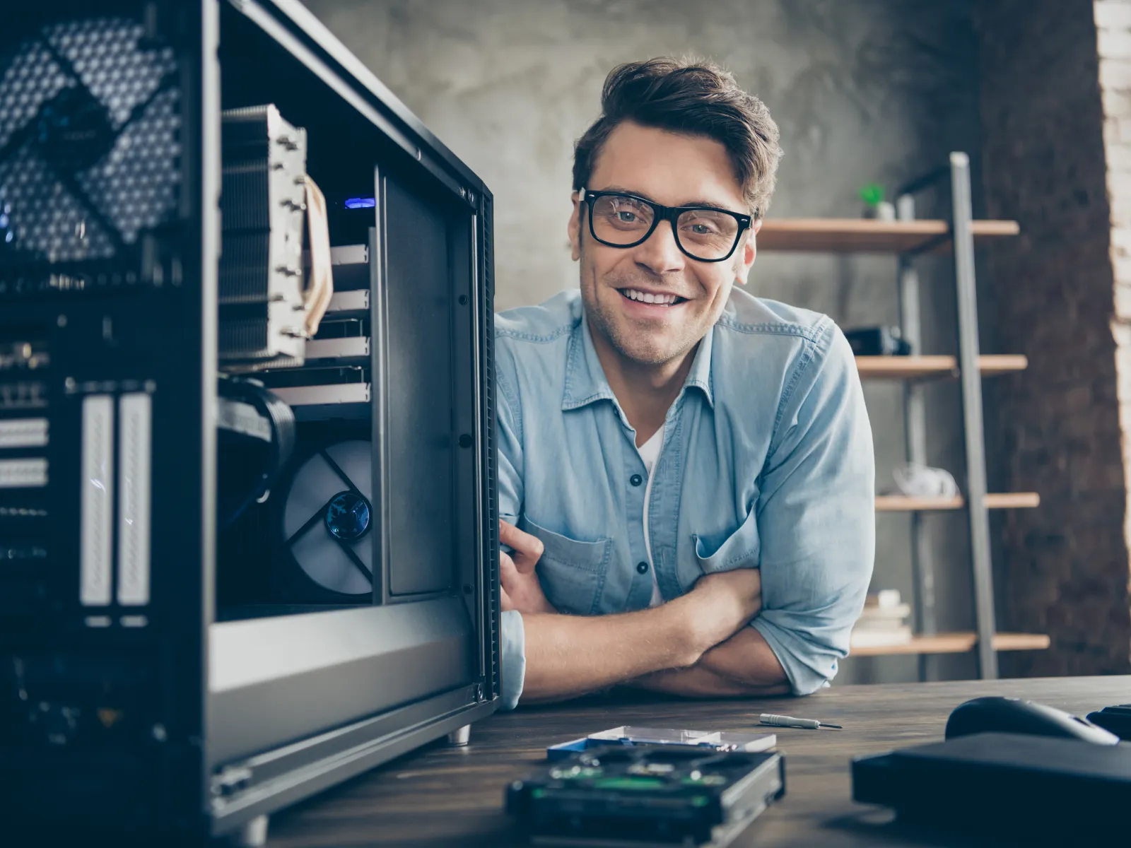 Man in glasses smiling next to an open desktop PC case with visible fans and components on desk.
