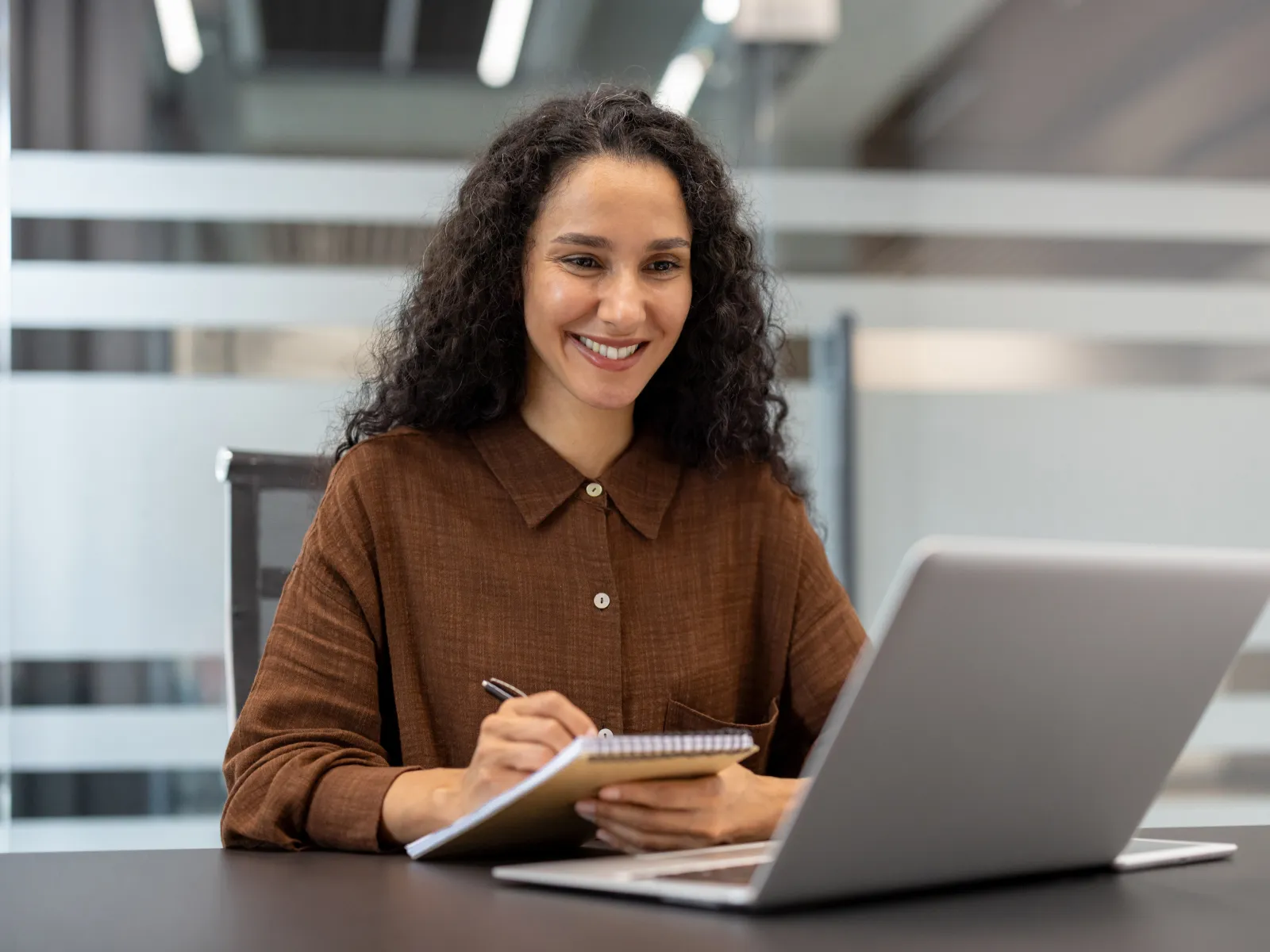 Smiling woman taking notes while looking at a laptop in a modern office environment.