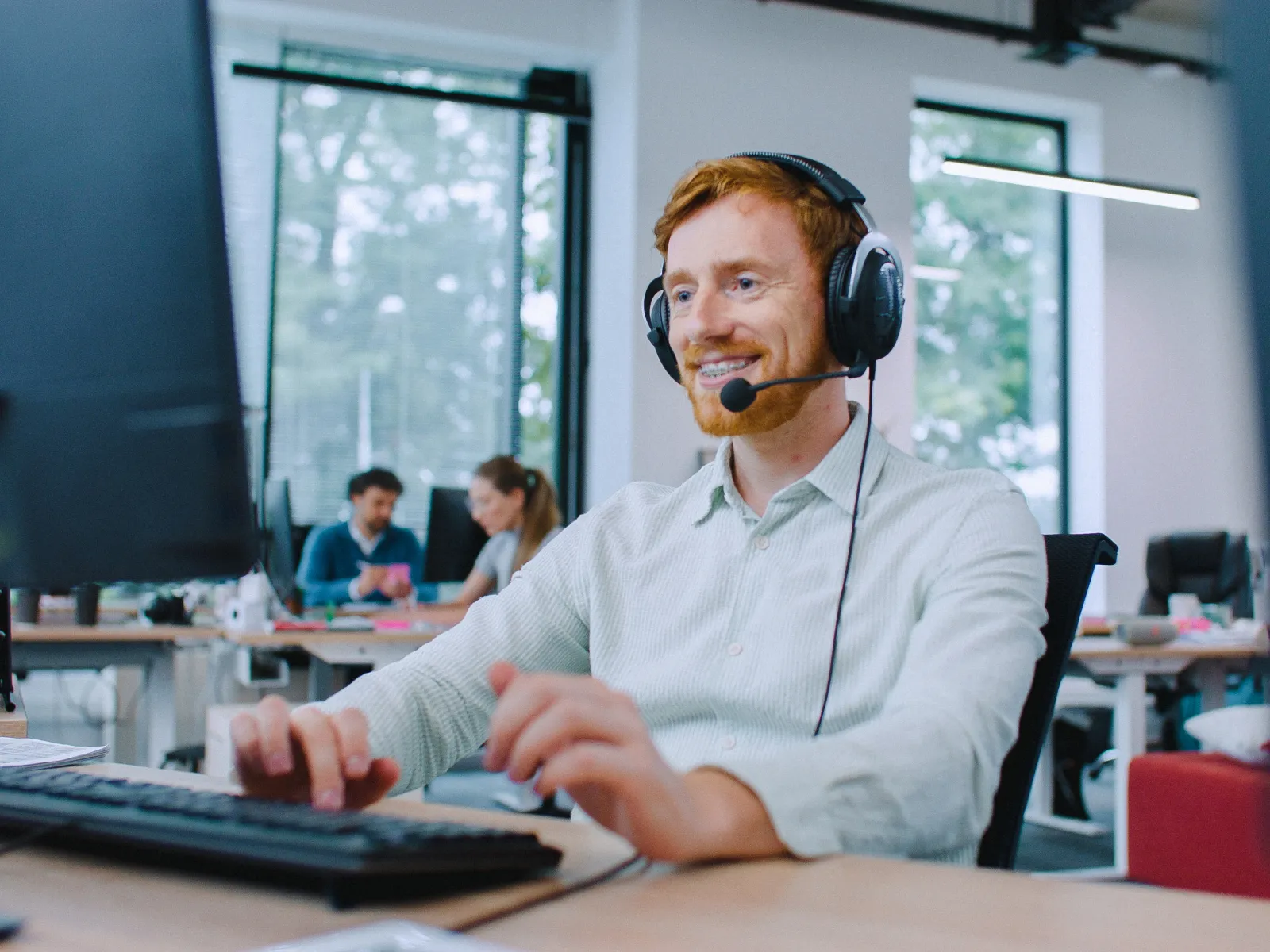 Happy man with headset working at computer in modern office with colleagues in the background.