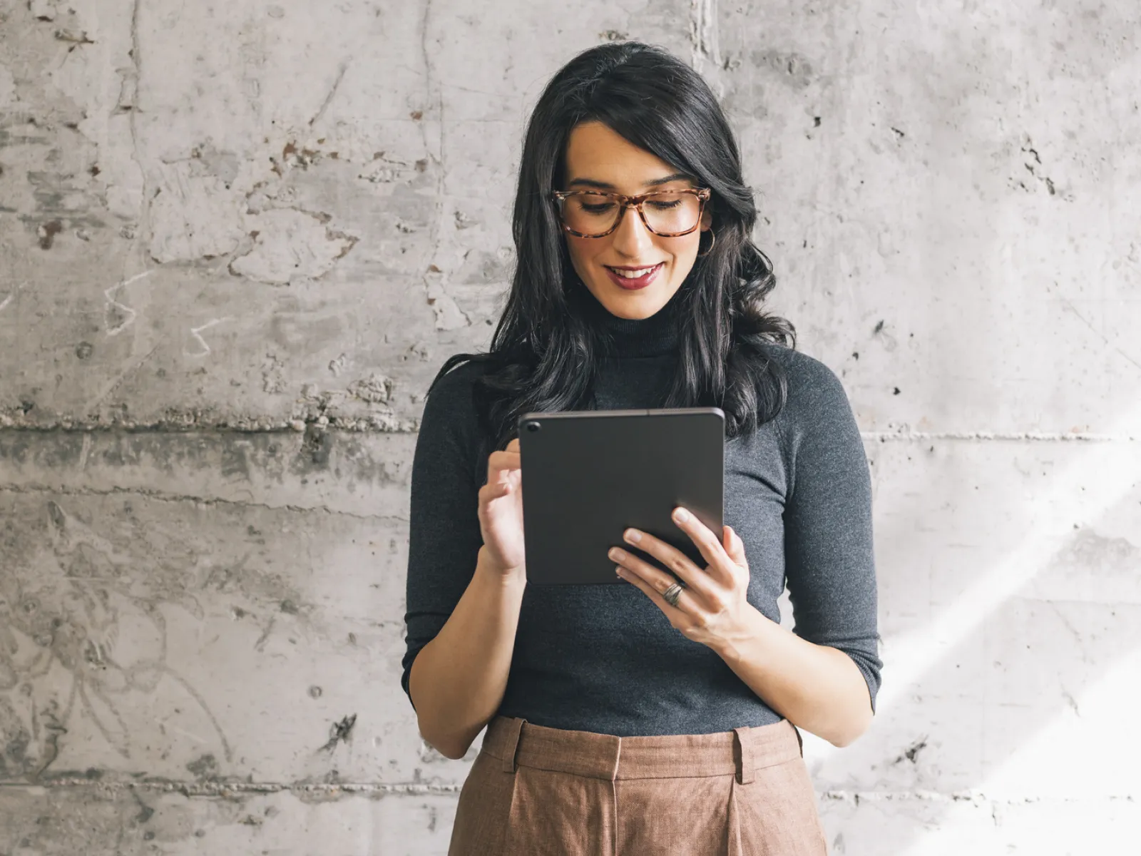 Woman with glasses using a tablet, smiling in a minimalistic gray setting.