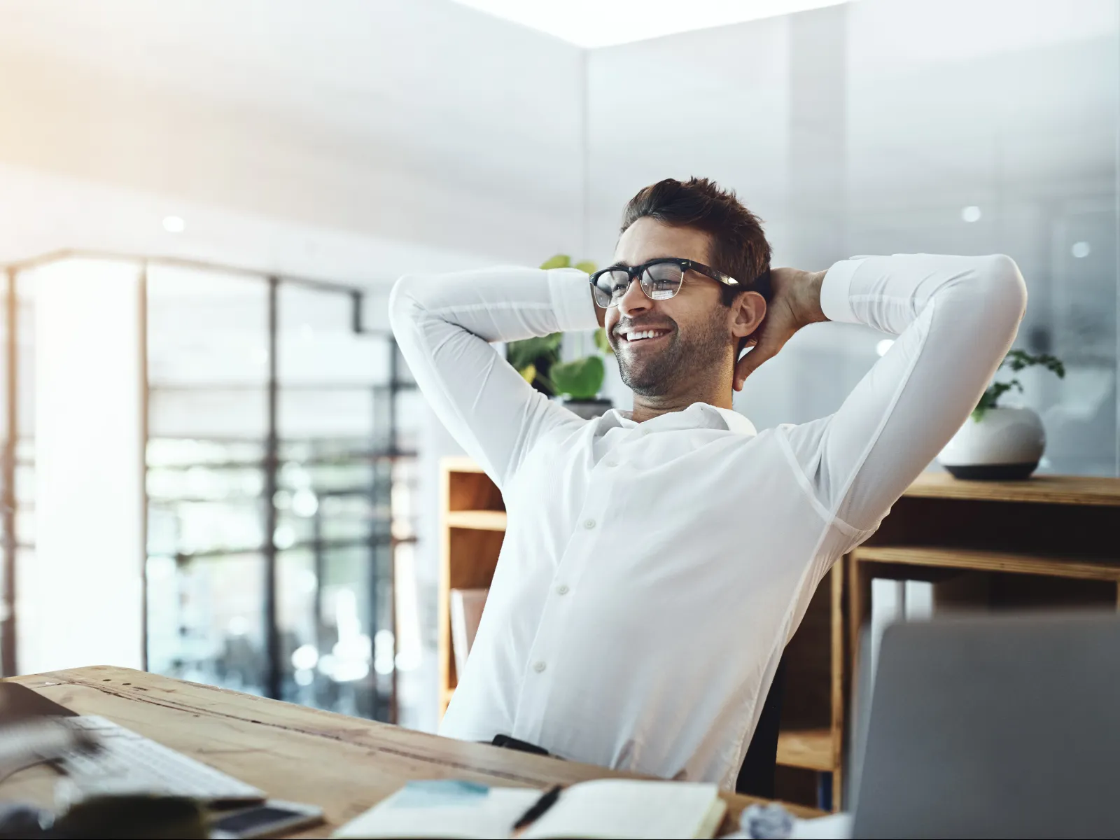 Smiling man in glasses relaxing at desk with hands behind head in bright modern office space.