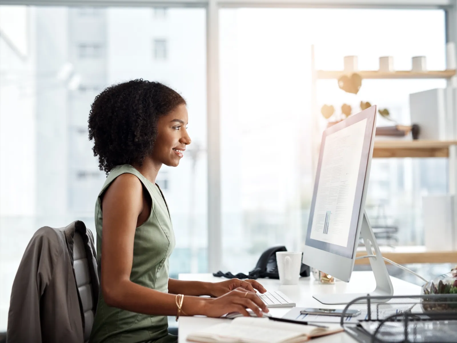 Young woman smiling and working on a desktop computer in a bright modern office with large windows.