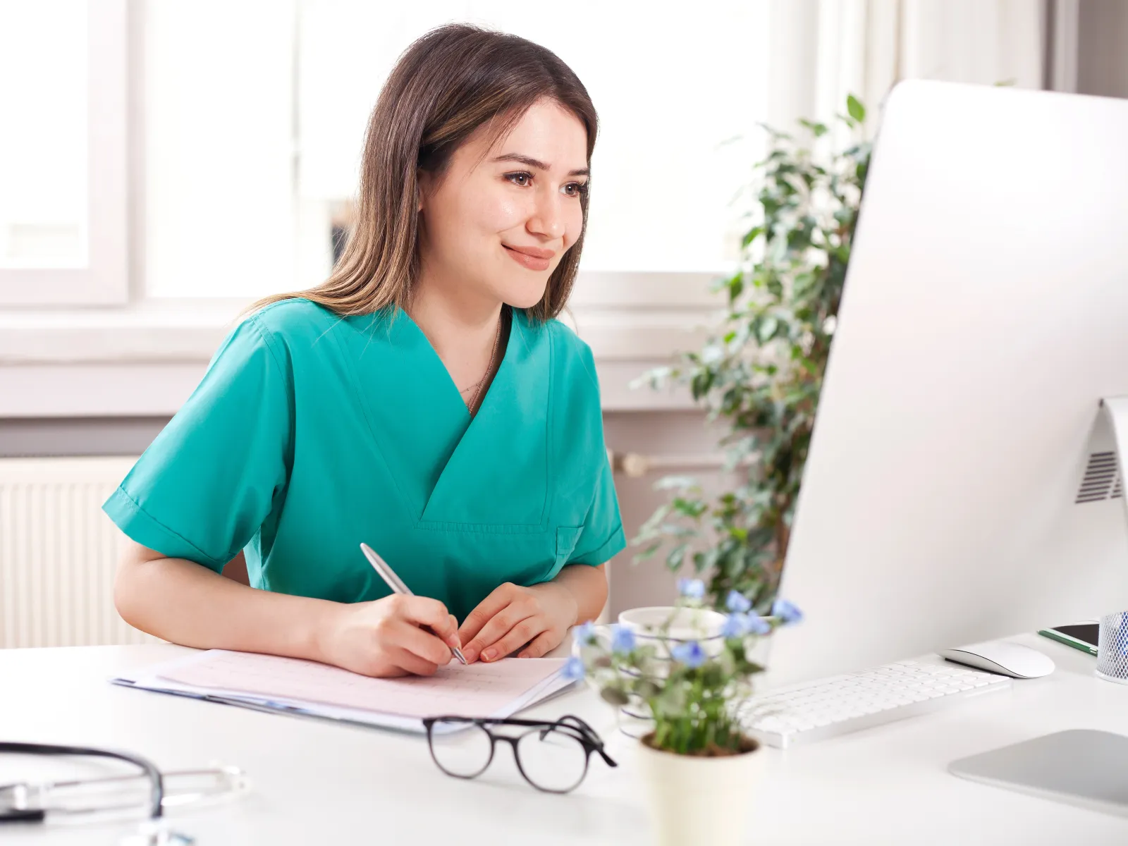 Female nurse in green scrubs smiling and writing notes at desk with computer and stethoscope nearby.