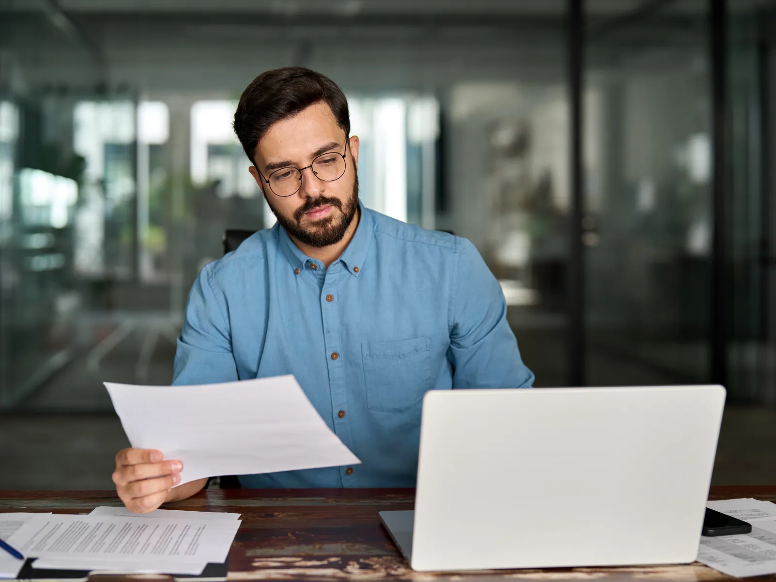 Man in glasses wearing blue shirt reviewing documents while working on a laptop at a wooden desk indoors.