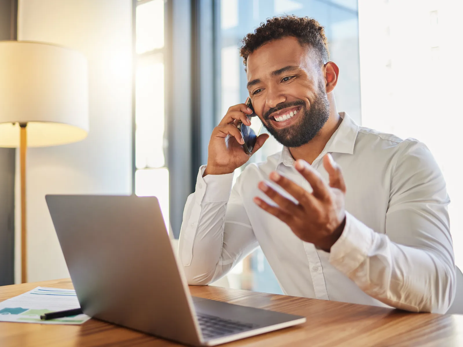 Smiling man in white shirt talking on phone while working on laptop in bright modern office.