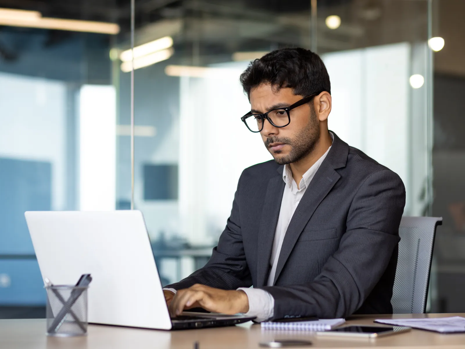 Young businessman in glasses working focused on laptop in modern office with glass walls and documents nearby