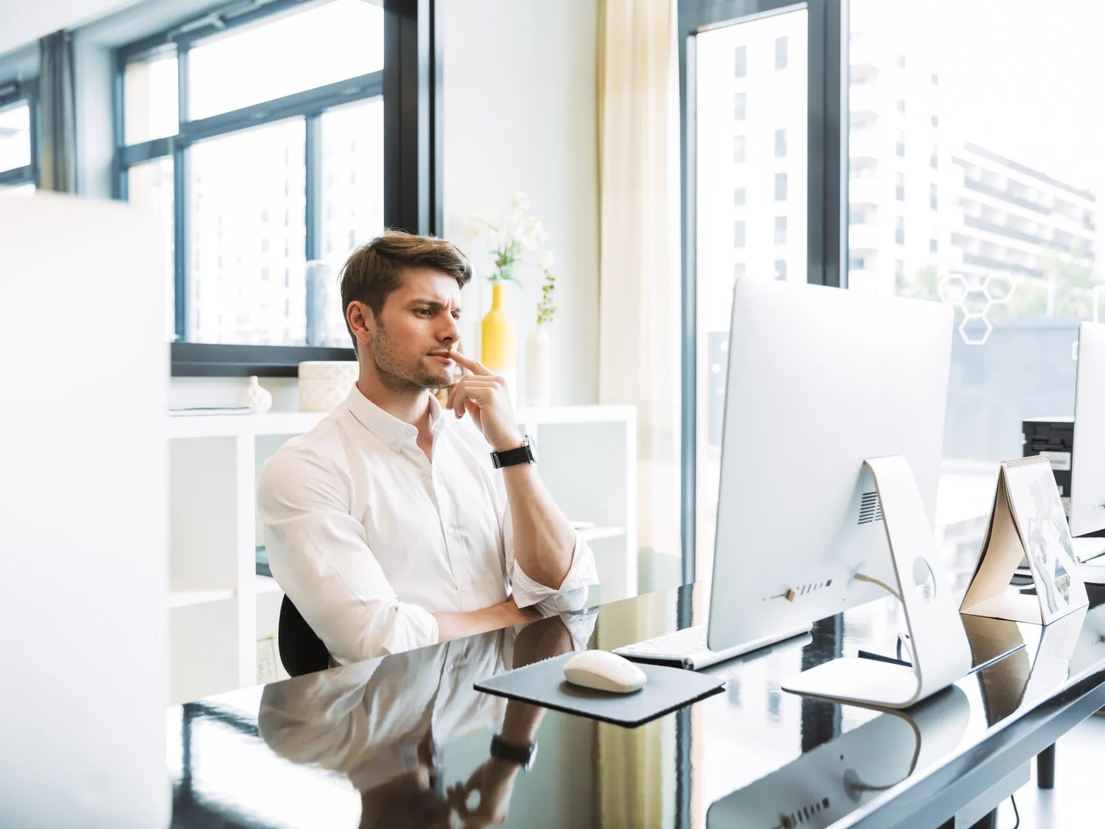 Man in white shirt thoughtfully working at a modern office desk with large computer screens and natural light.
