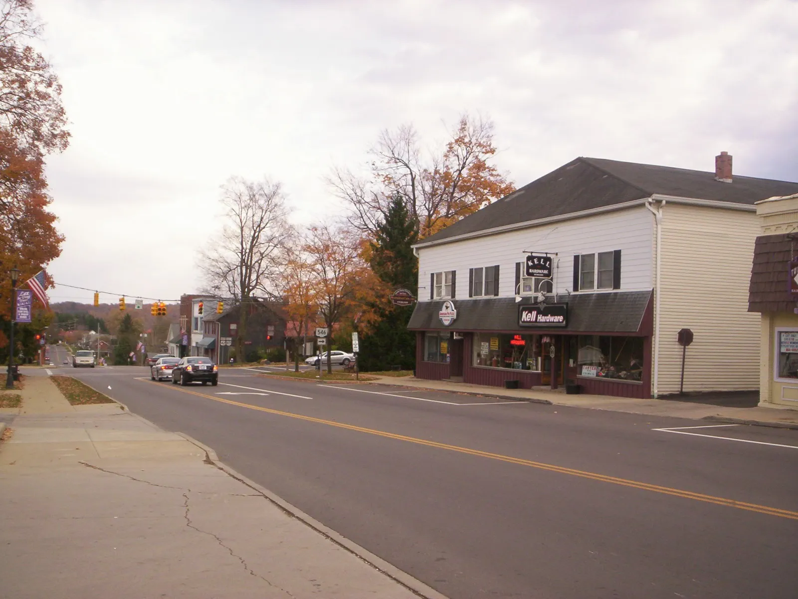 Quiet small town street with a hardware store, fall trees, and light traffic under an overcast sky