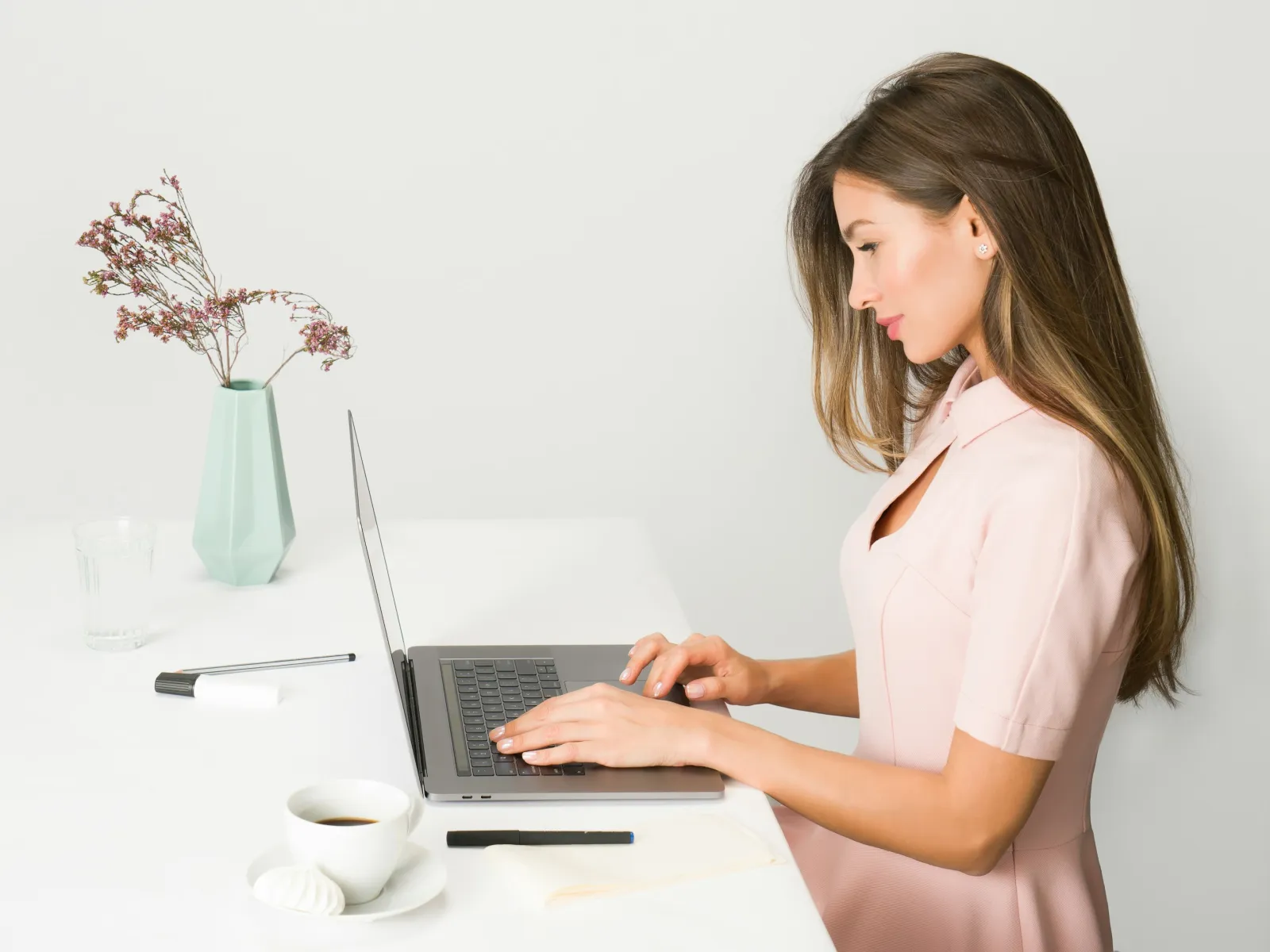 Young woman in pink dress typing on laptop at white desk with coffee and vase with flowers.