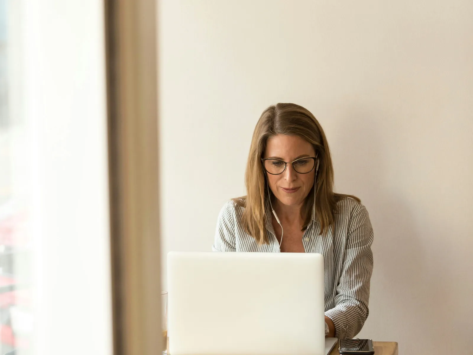 Woman wearing glasses working on laptop at wooden table with newspaper and phone in cozy indoor space.