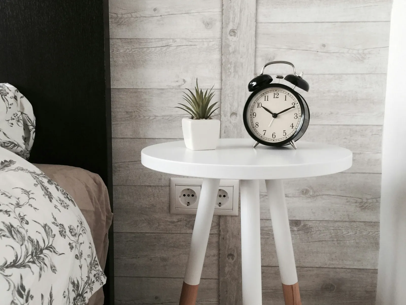 Modern bedside table with a small plant and retro black alarm clock in a cozy bedroom setting.