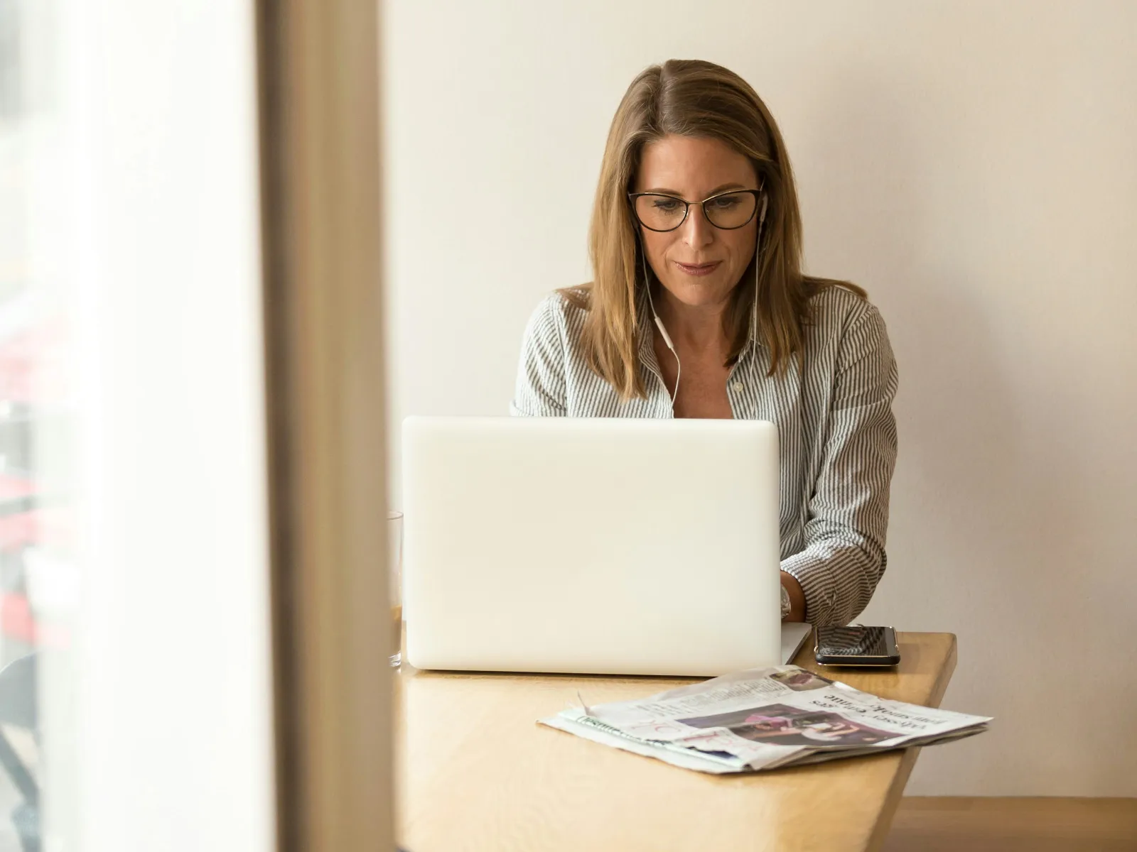 Woman with glasses using a laptop at a wooden table with newspapers and phone nearby in a bright room.