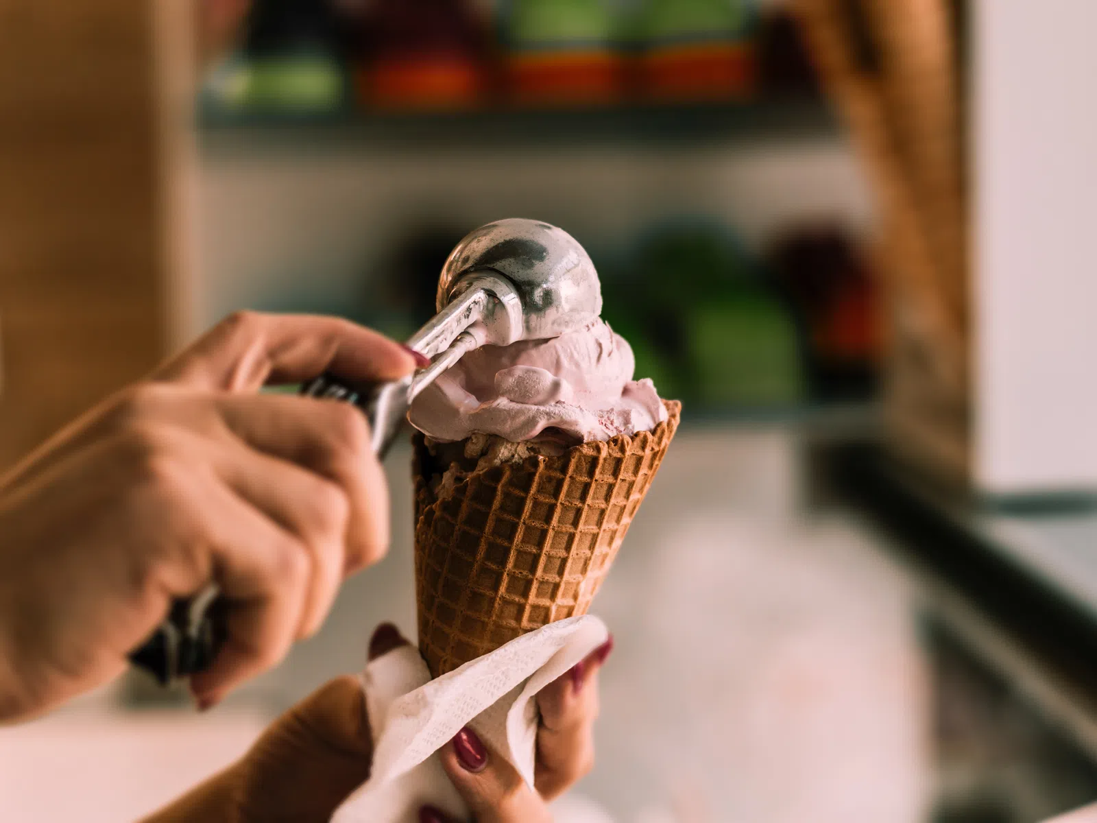 Hand scooping pink ice cream onto a waffle cone in a shop with blurred background.