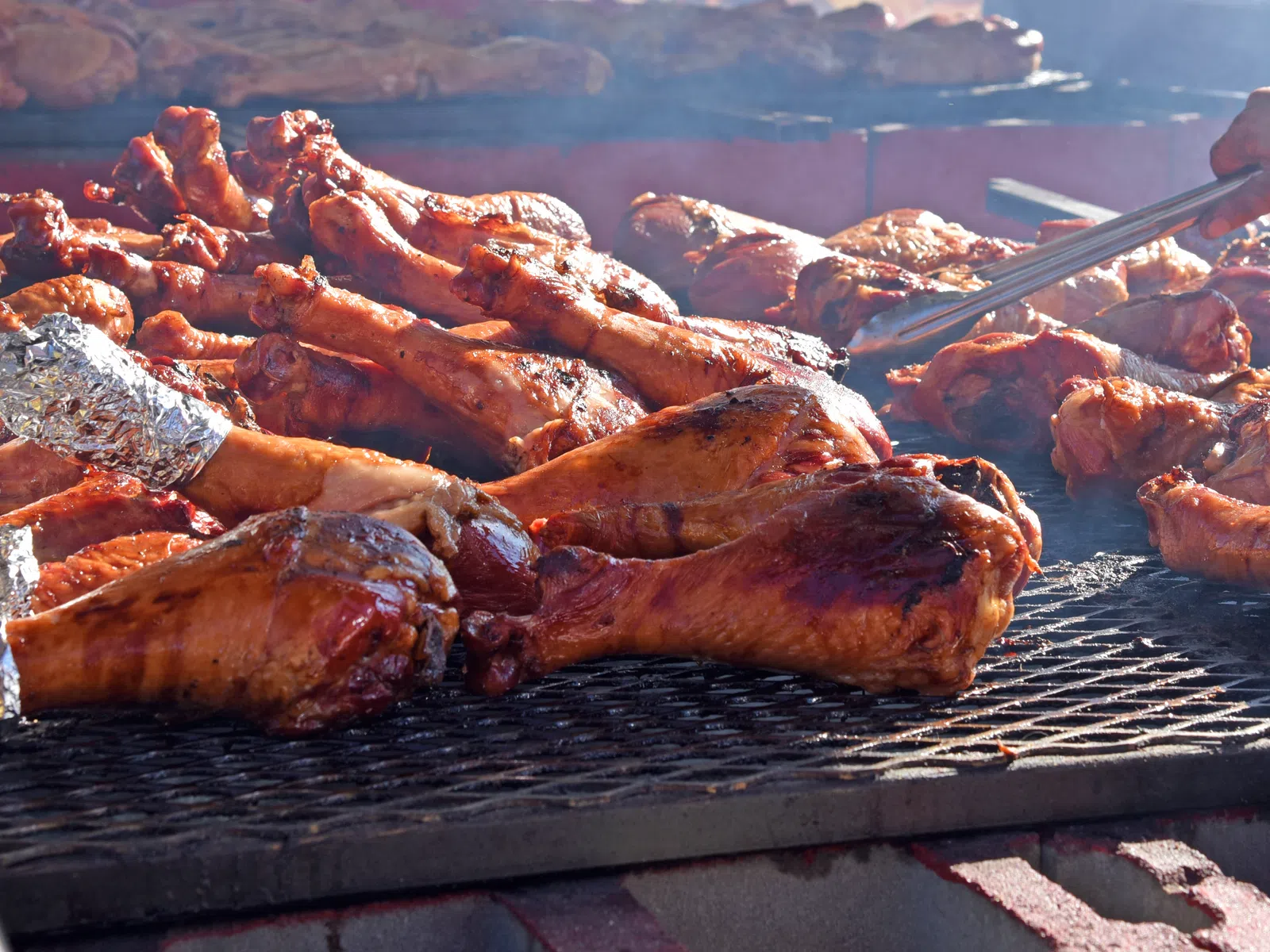 Grilled turkey legs cooking over a charcoal barbecue with hands turning meat using tongs on an outdoor grill.
