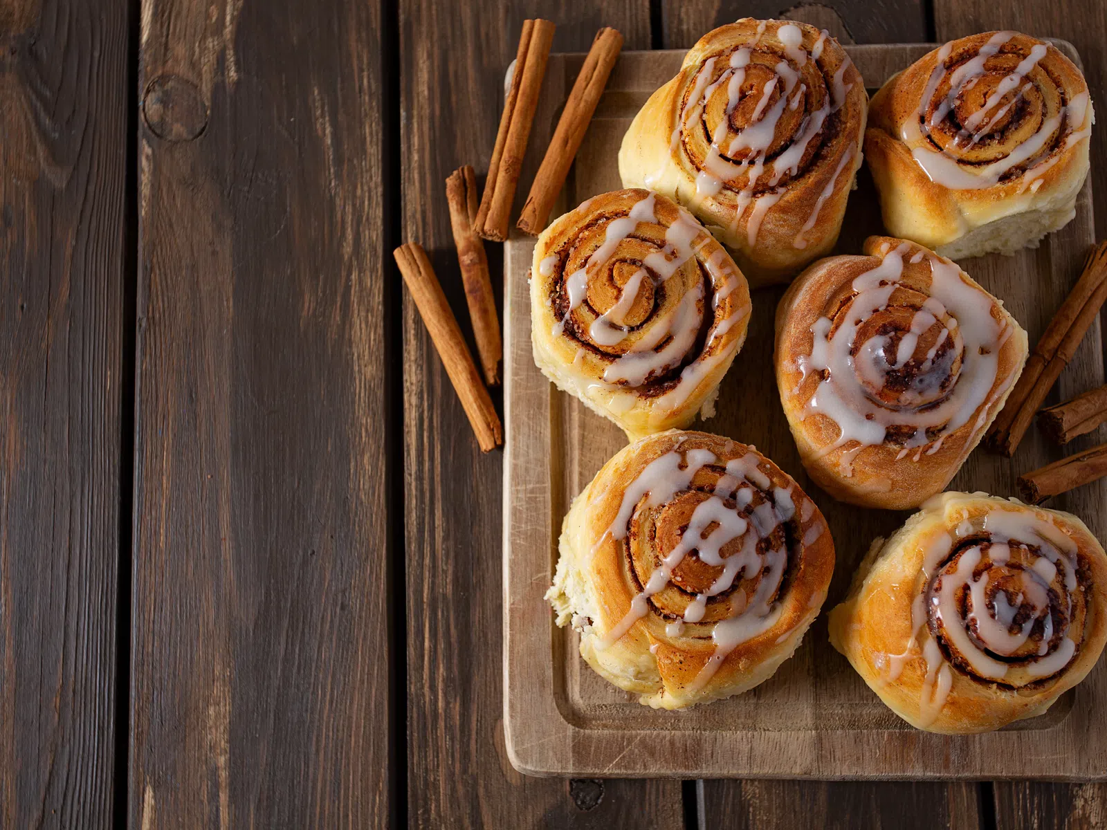 Fresh cinnamon rolls with icing on a wooden cutting board surrounded by cinnamon sticks on dark wood table.