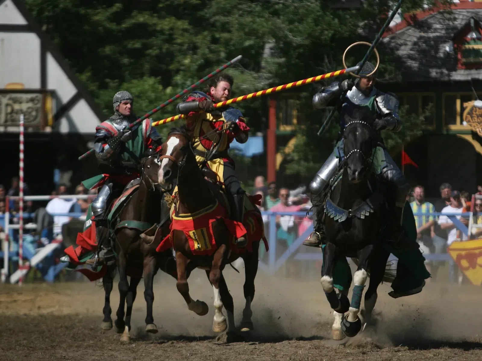 Three armored knights jousting on horses at a medieval festival with crowd and banners in the background.