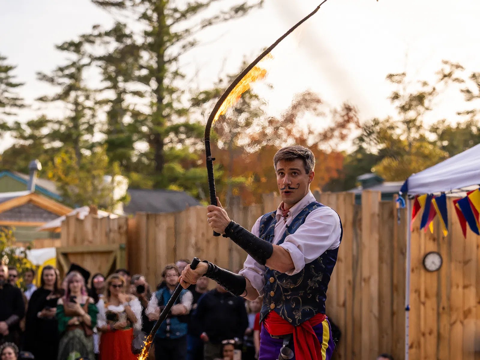 Fire performer in colorful medieval costume twirling a flaming whip on outdoor stage at festival with audience.