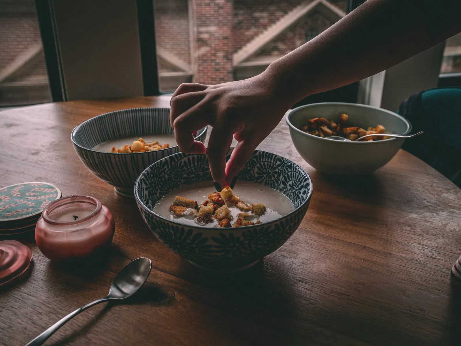 Hand placing cereal into a decorative bowl of milk on a wooden table with two other bowls in the background