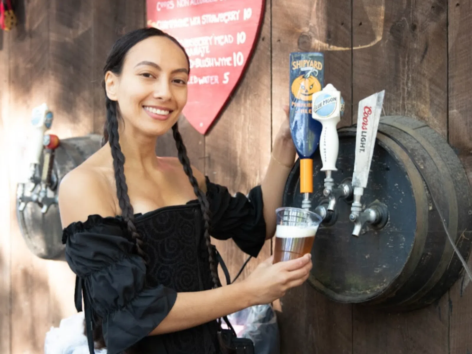 Smiling woman in black dress pouring beer from a tap attached to a wooden barrel outdoors.
