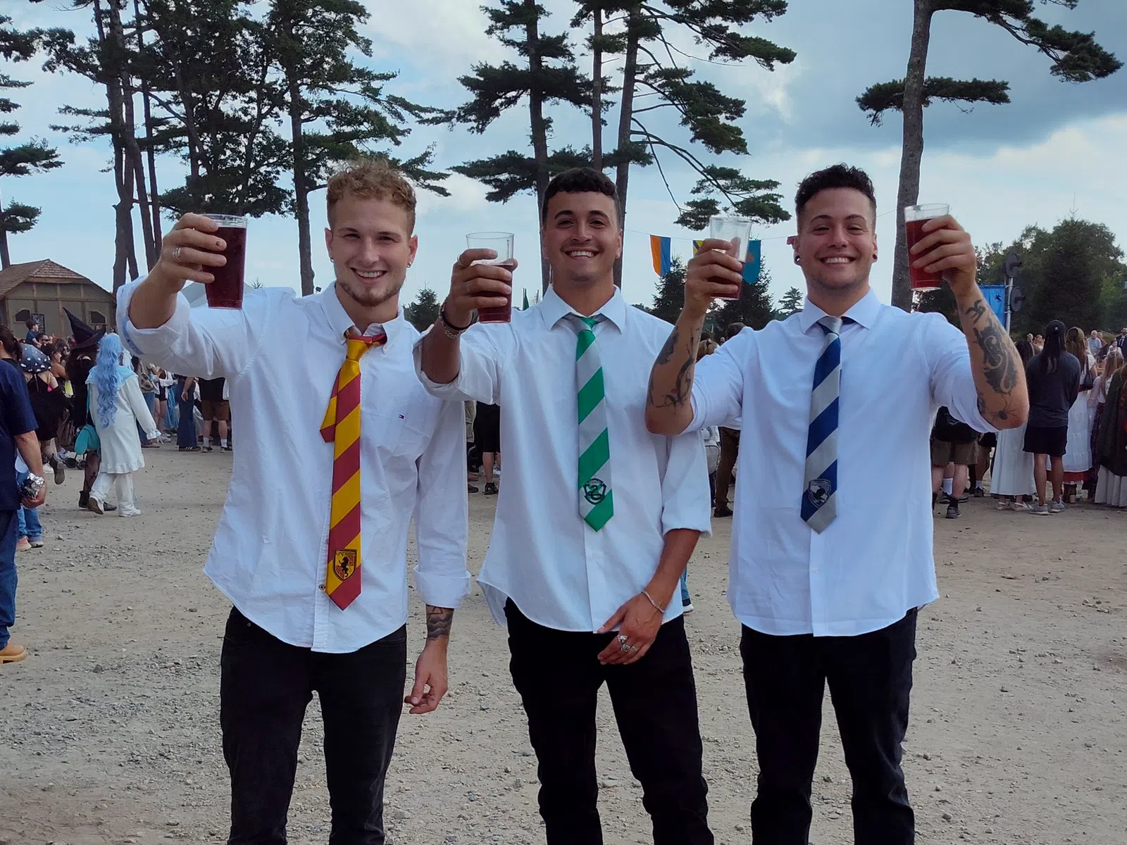 Three young men in white shirts and colorful ties holding drinks and smiling outdoors with trees in background.