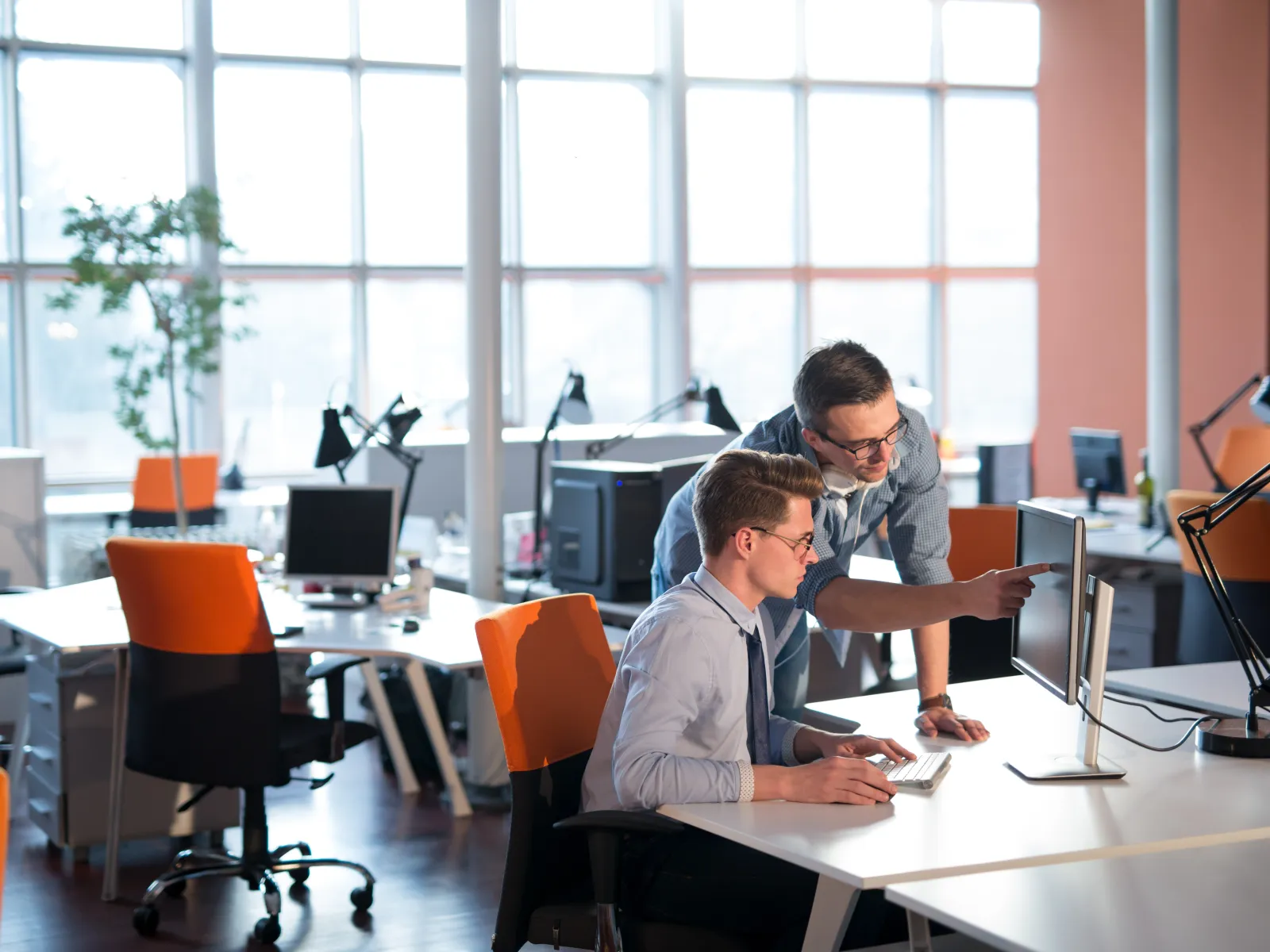 Two coworkers collaborate on a desktop computer in a modern office with large windows and orange chairs.