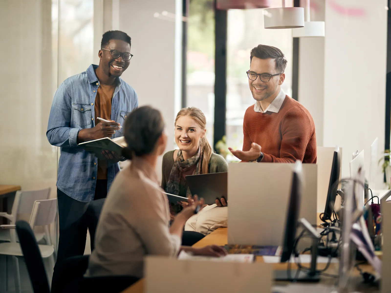 Four diverse coworkers smiling and collaborating in a modern office with computers and large windows.