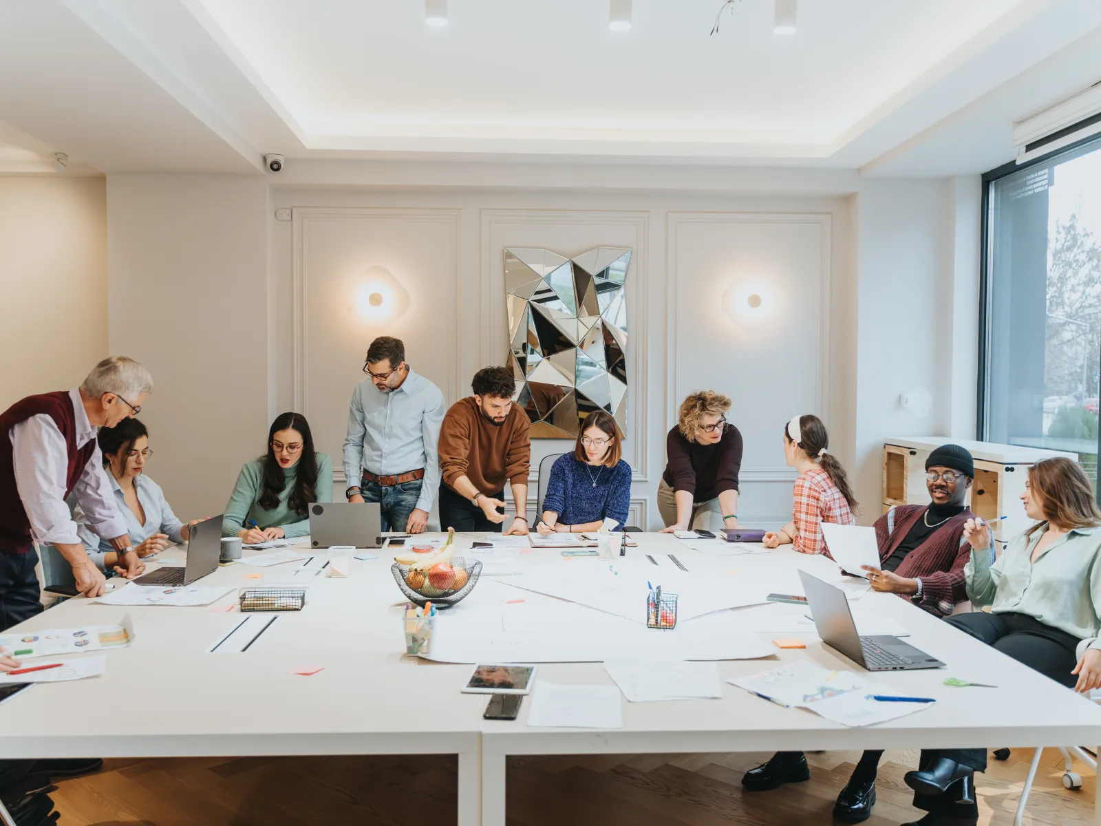 Diverse team collaborating around a large white table with laptops and documents in a modern office.