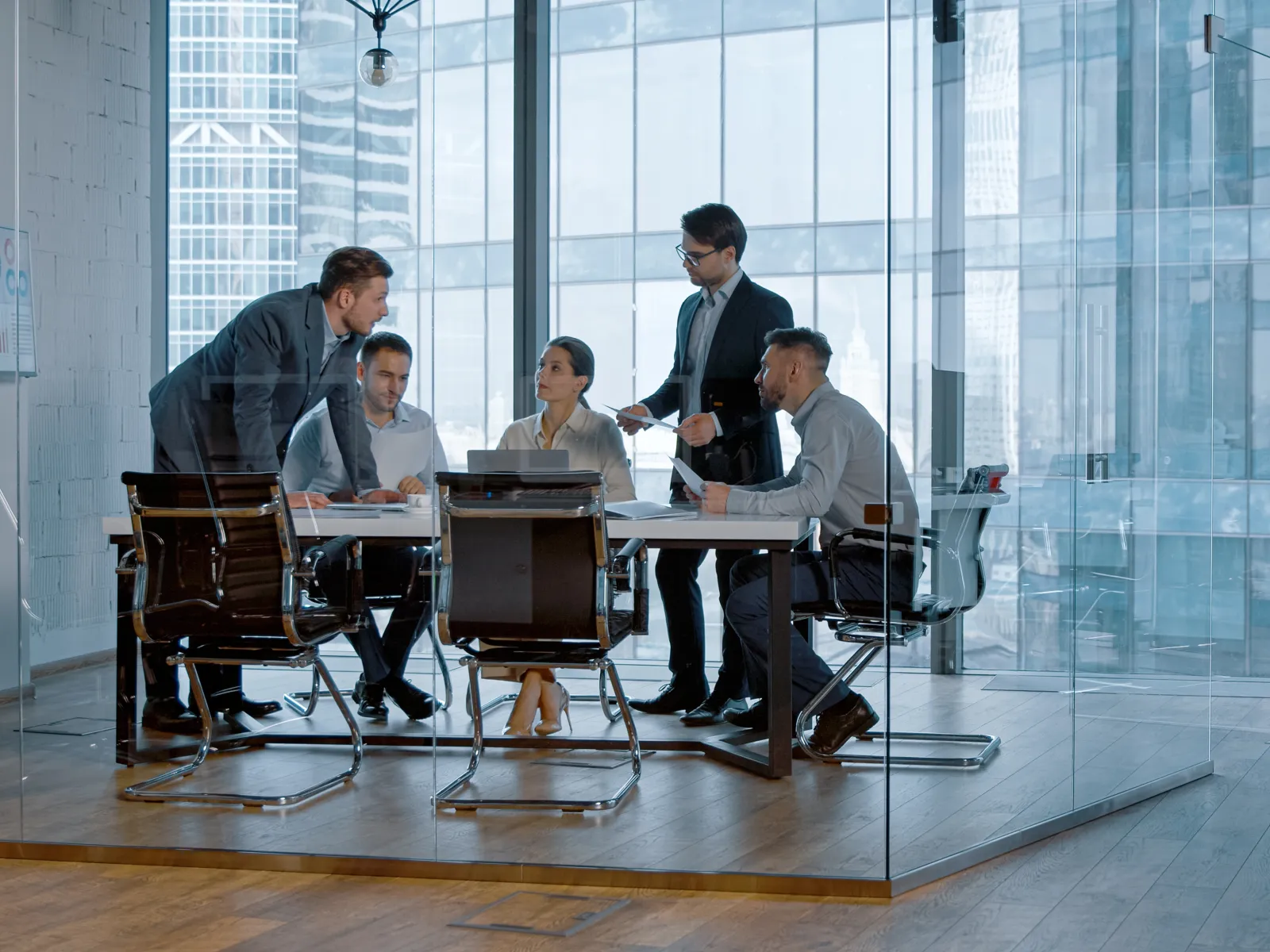 Five professionals in formal attire engaged in a meeting around a conference table in a modern glass office.