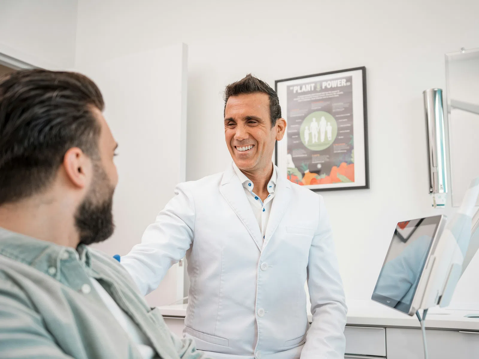 Smiling doctor in white coat talking to male patient in modern medical office with computer and plant poster.