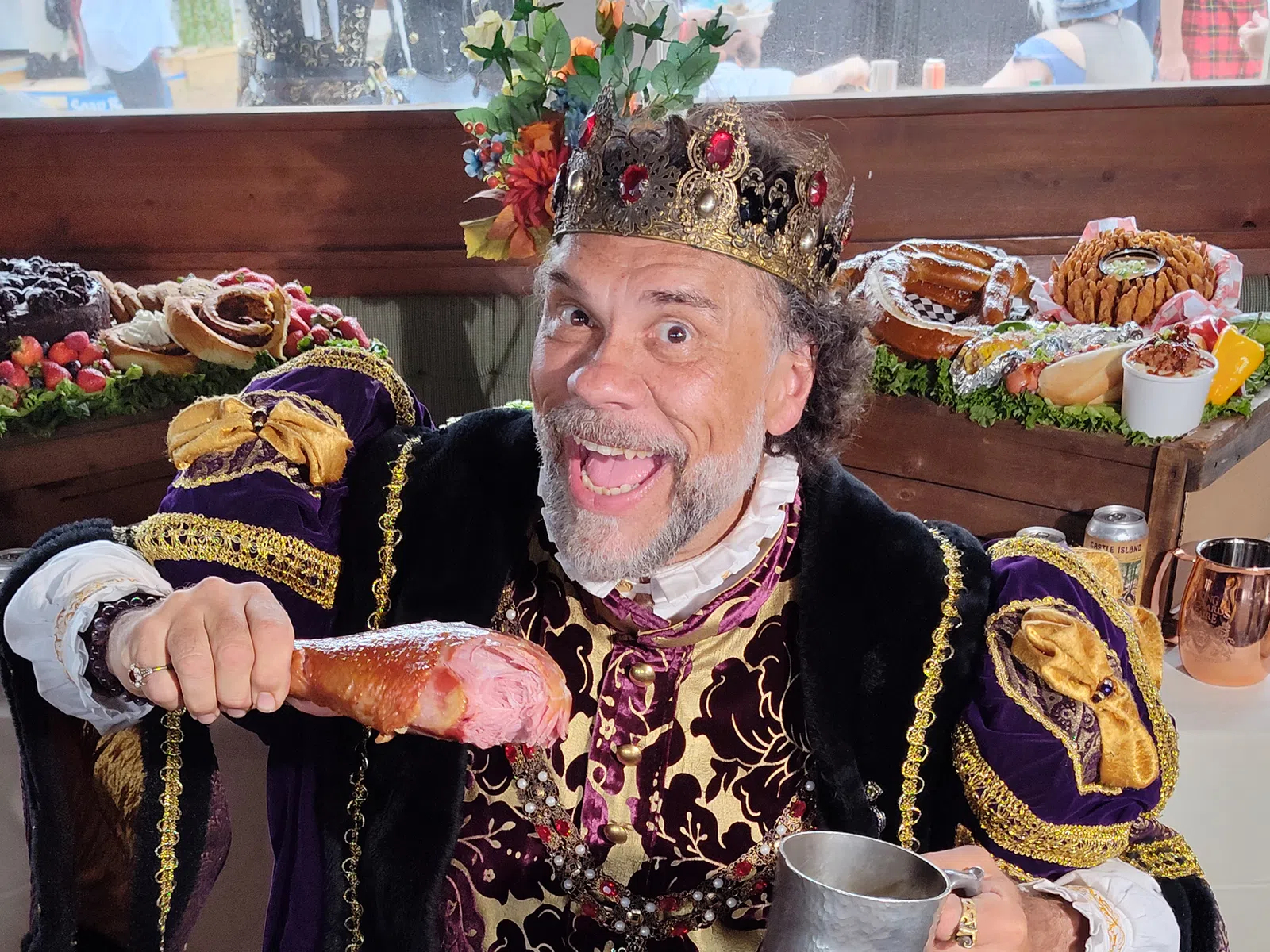 Three people in medieval attire serve food including roasted turkey legs under a blue and purple tent at an outdoor event.