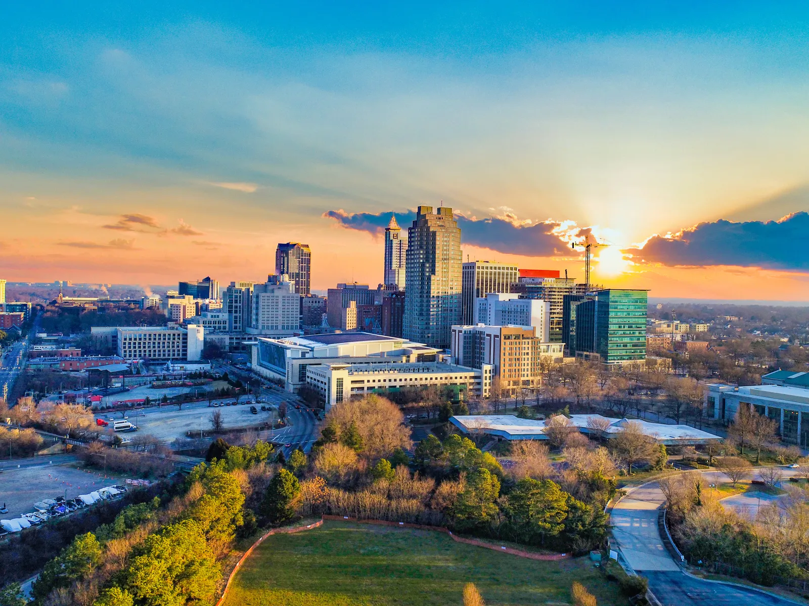 Sunset view over a city skyline with tall buildings and green parkland in the foreground.