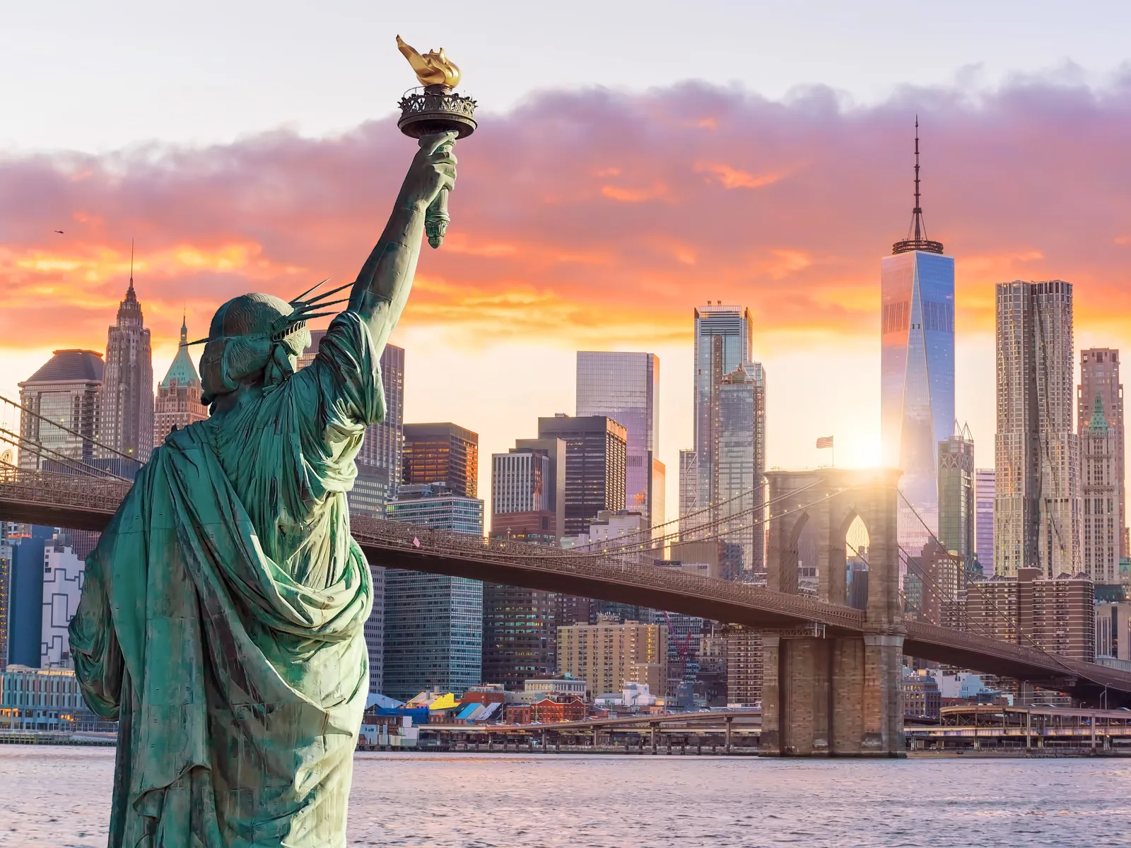 Sunset view of Statue of Liberty with Brooklyn Bridge and New York City skyline in the background