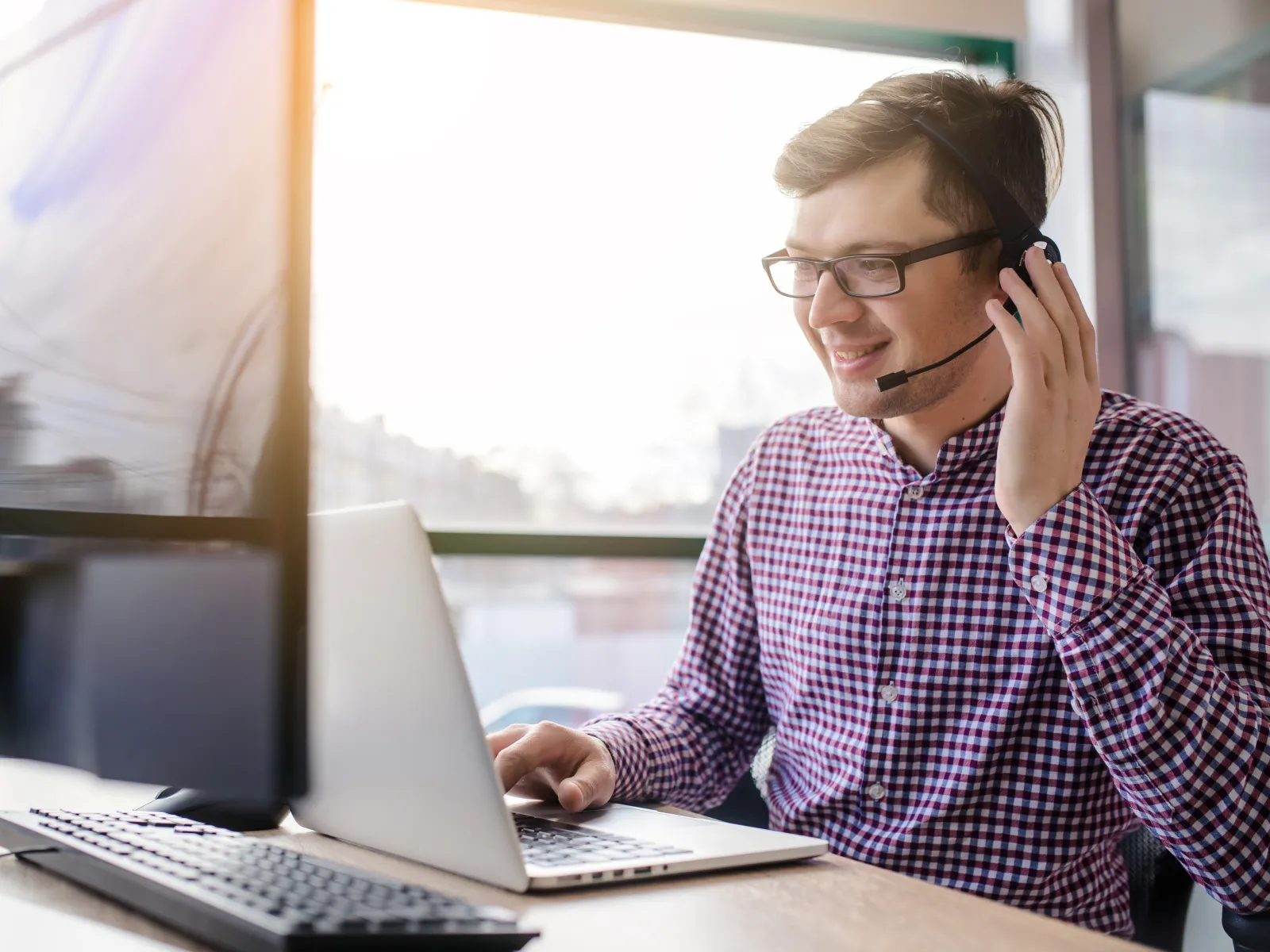 Smiling man in headset working on laptop in a bright office environment.