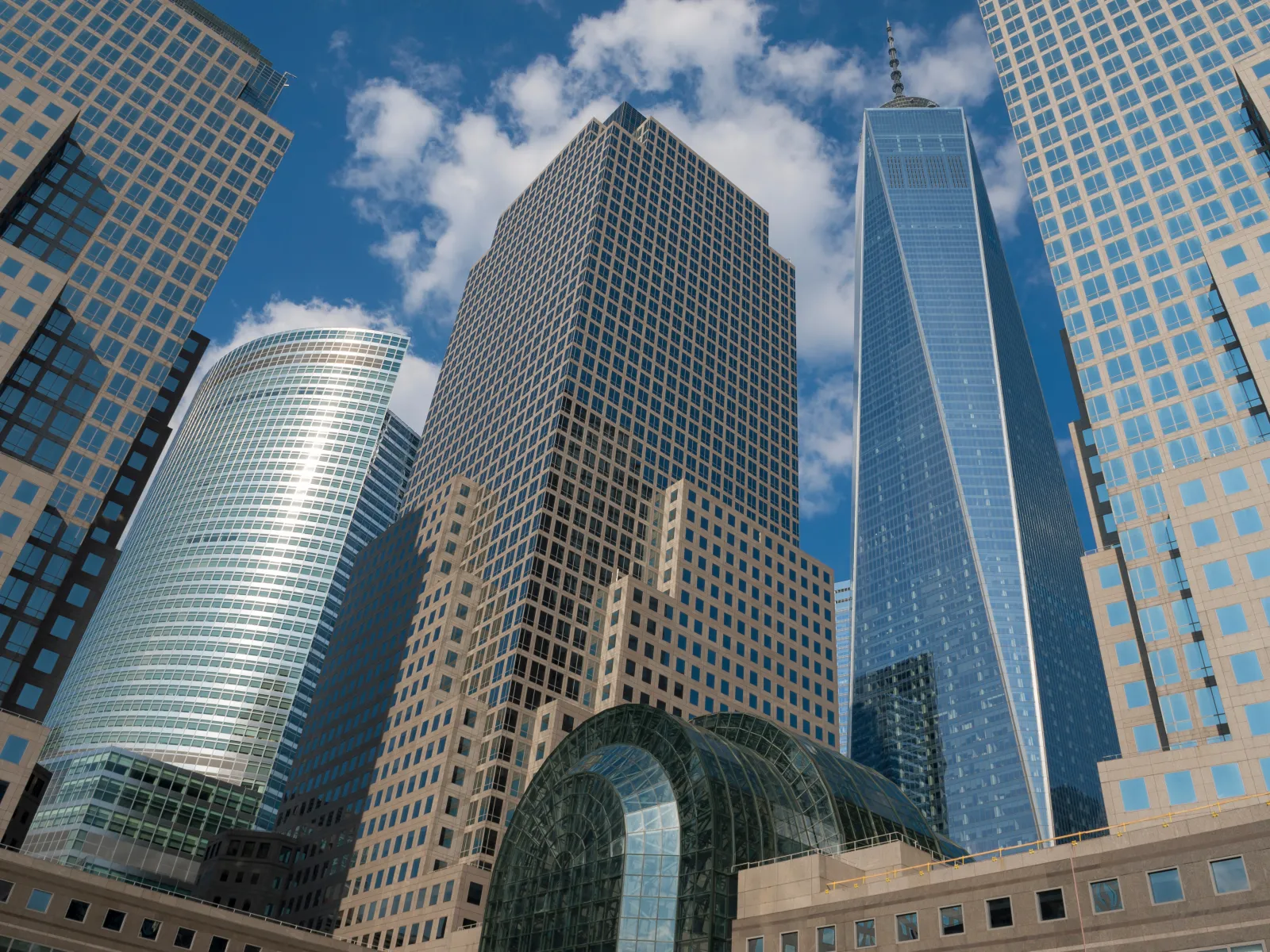 Modern skyscrapers including One World Trade Center under a partly cloudy blue sky in a cityscape.