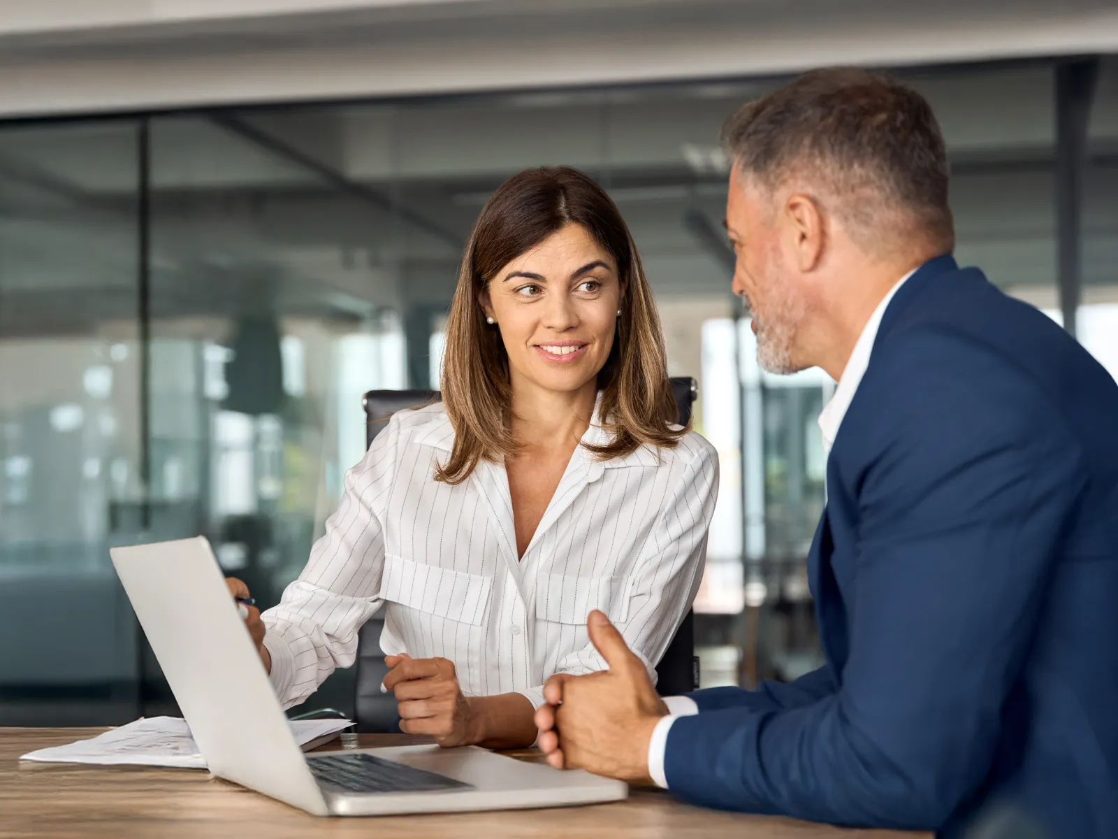 Two professionals discussing business at a modern office desk with a laptop and documents.