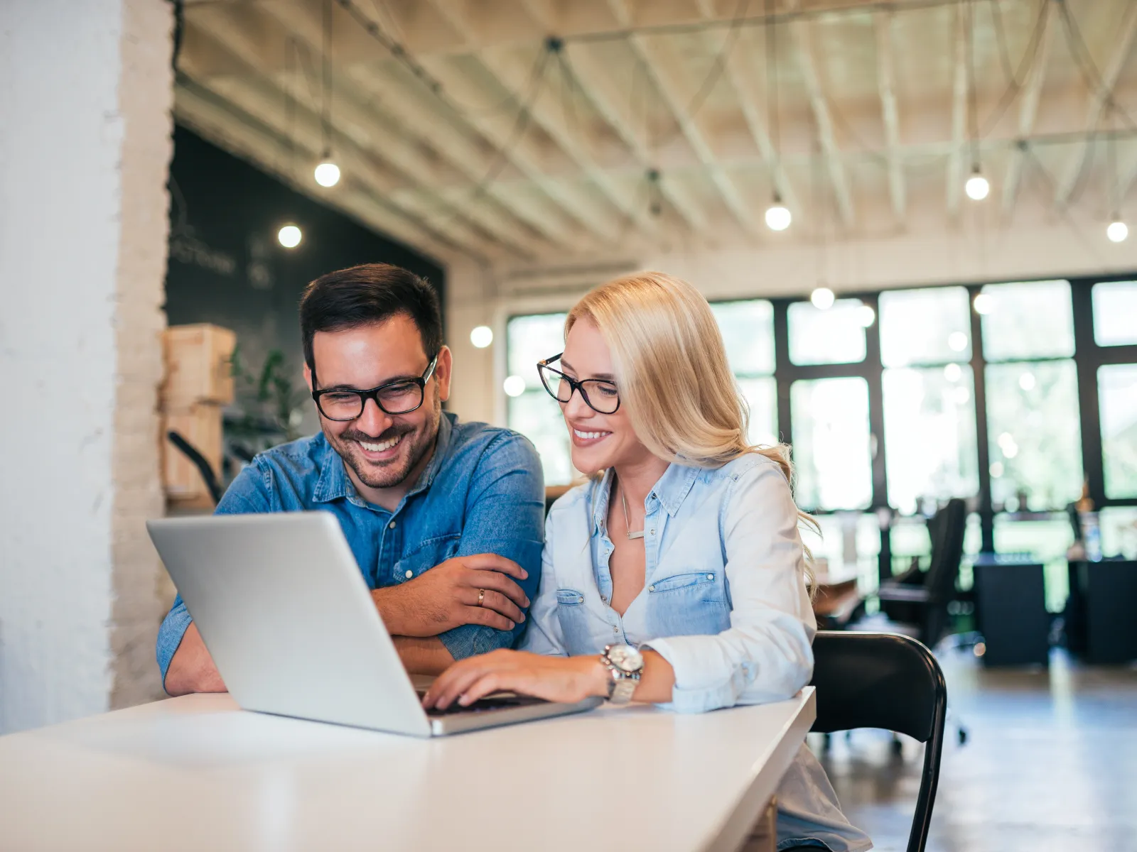 Smiling man and woman wearing glasses working together on a laptop in a bright modern office space.