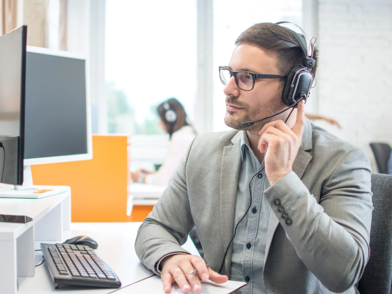 Man wearing headset and glasses working at computer in modern office call center environment.