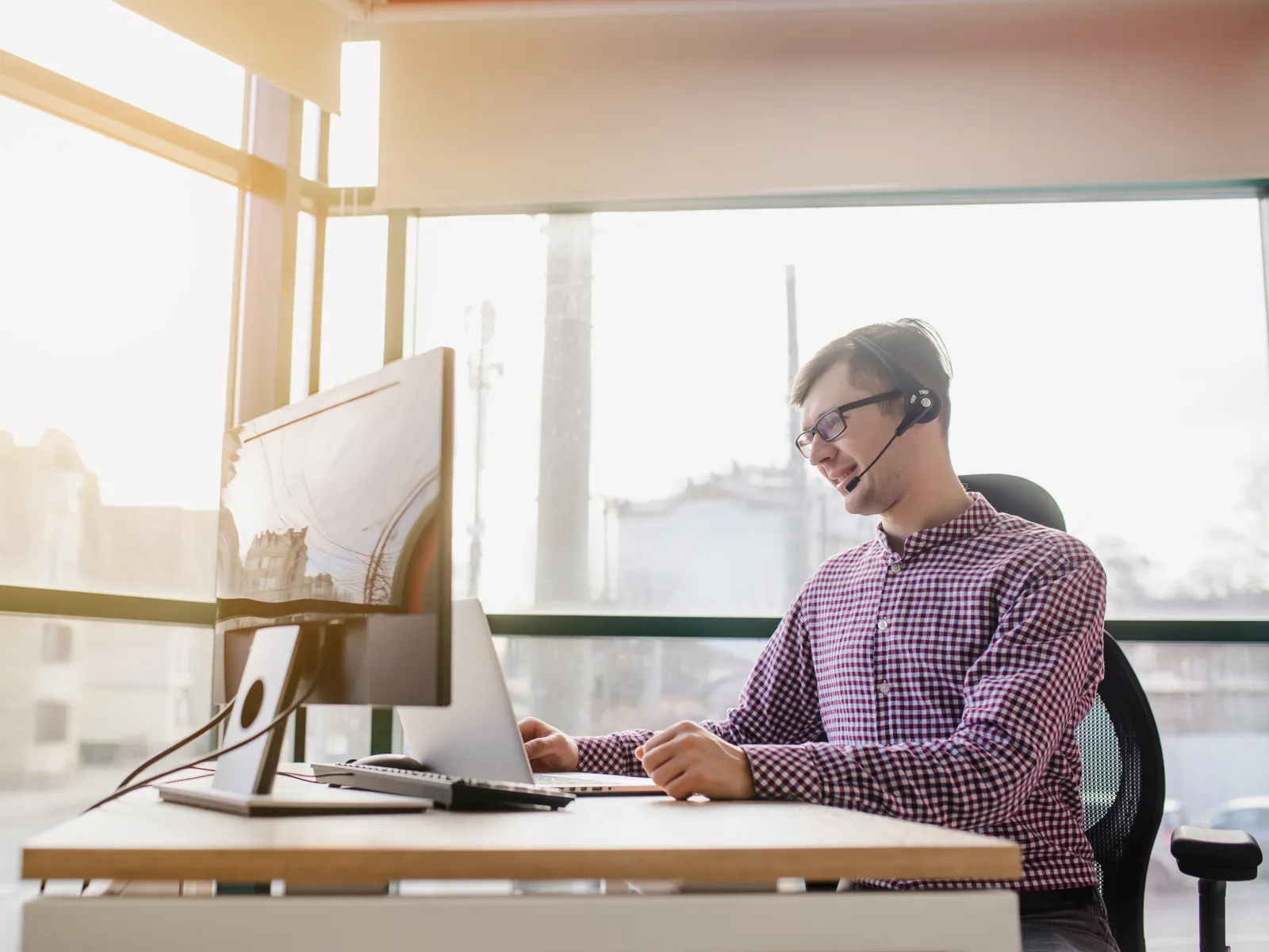 Man with headset working on computer in bright office with large windows and natural light.
