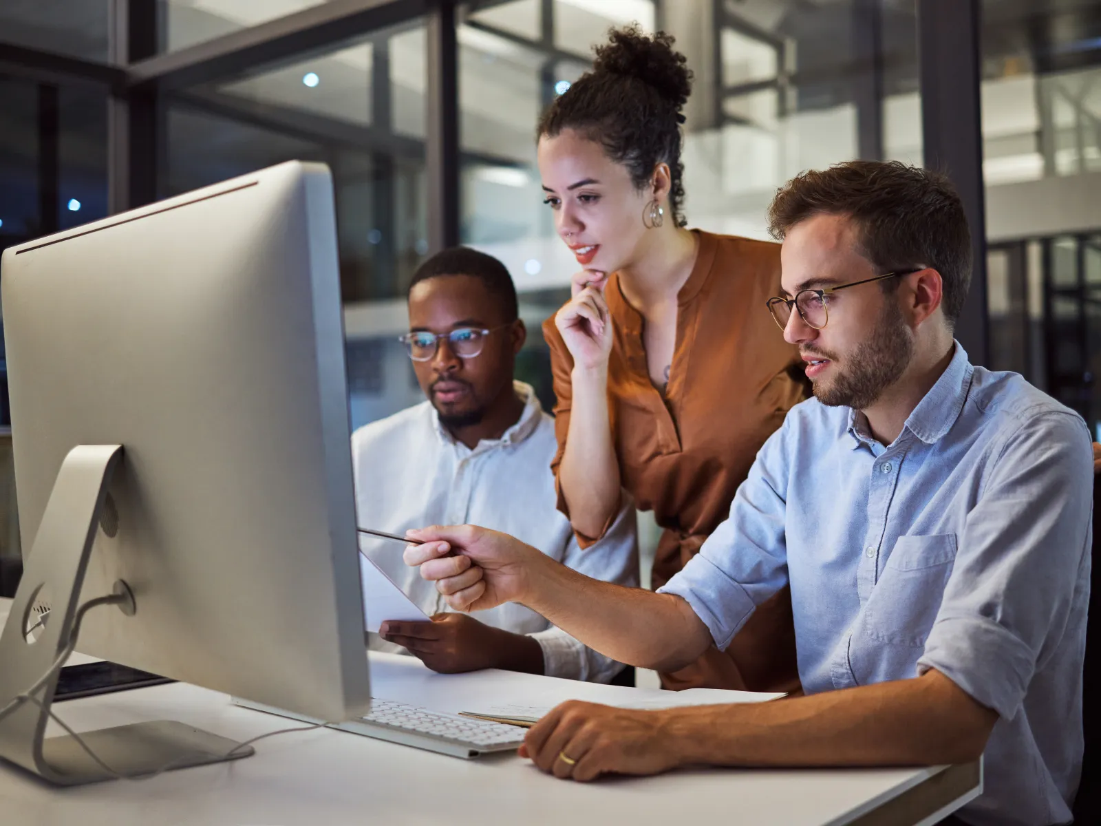 Three diverse colleagues collaborating and discussing work on a large desktop computer in a modern office.