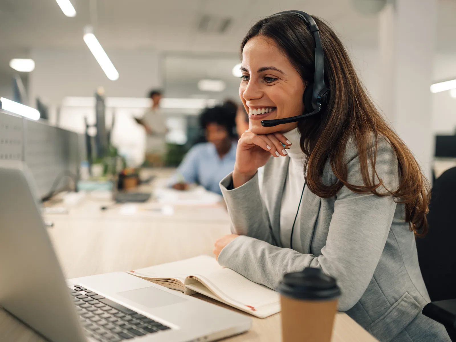Smiling woman wearing headset working on laptop in modern office with coffee and open notebook