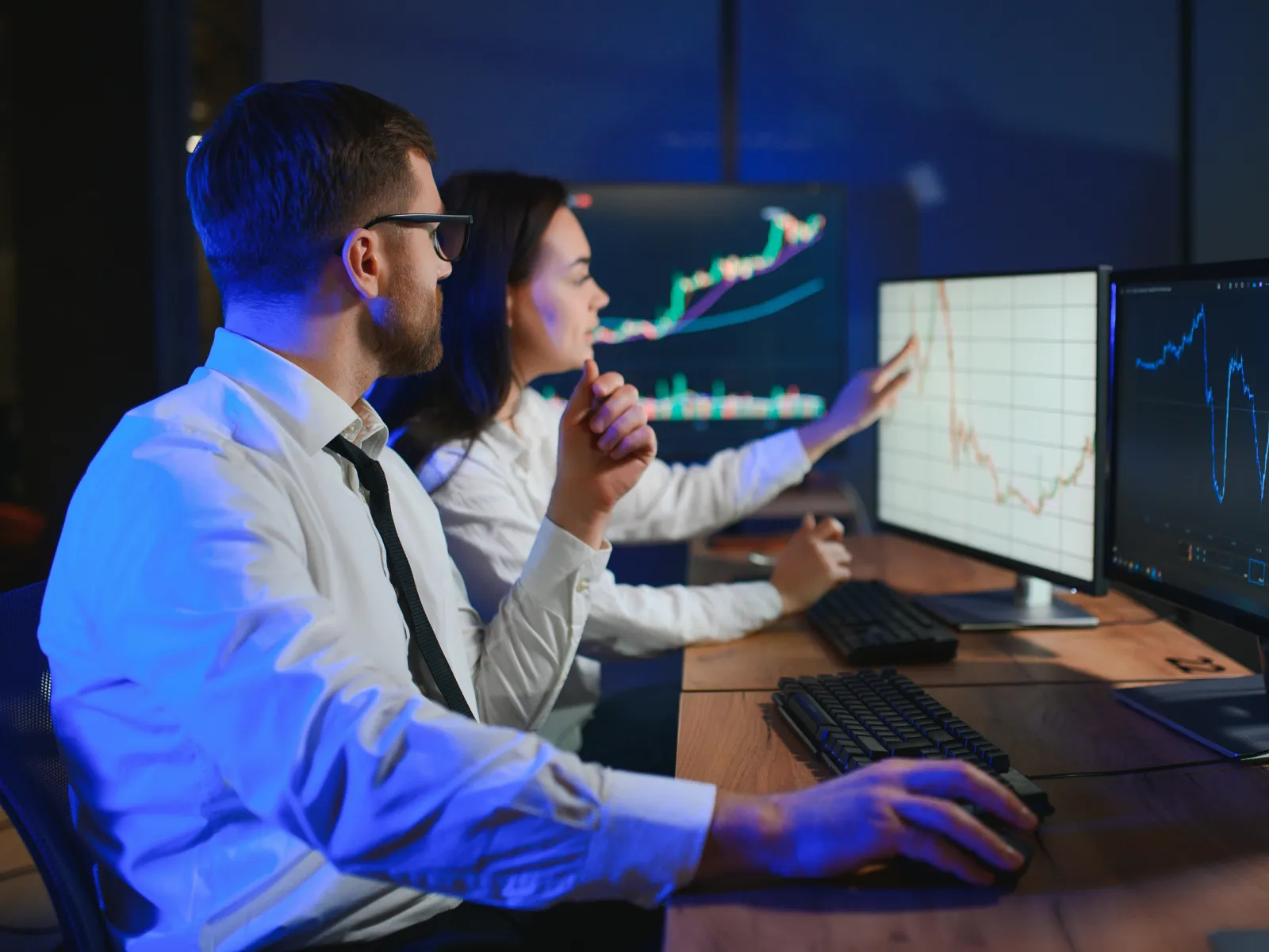 Two analysts in office reviewing stock market charts on multiple computer screens during night work session.