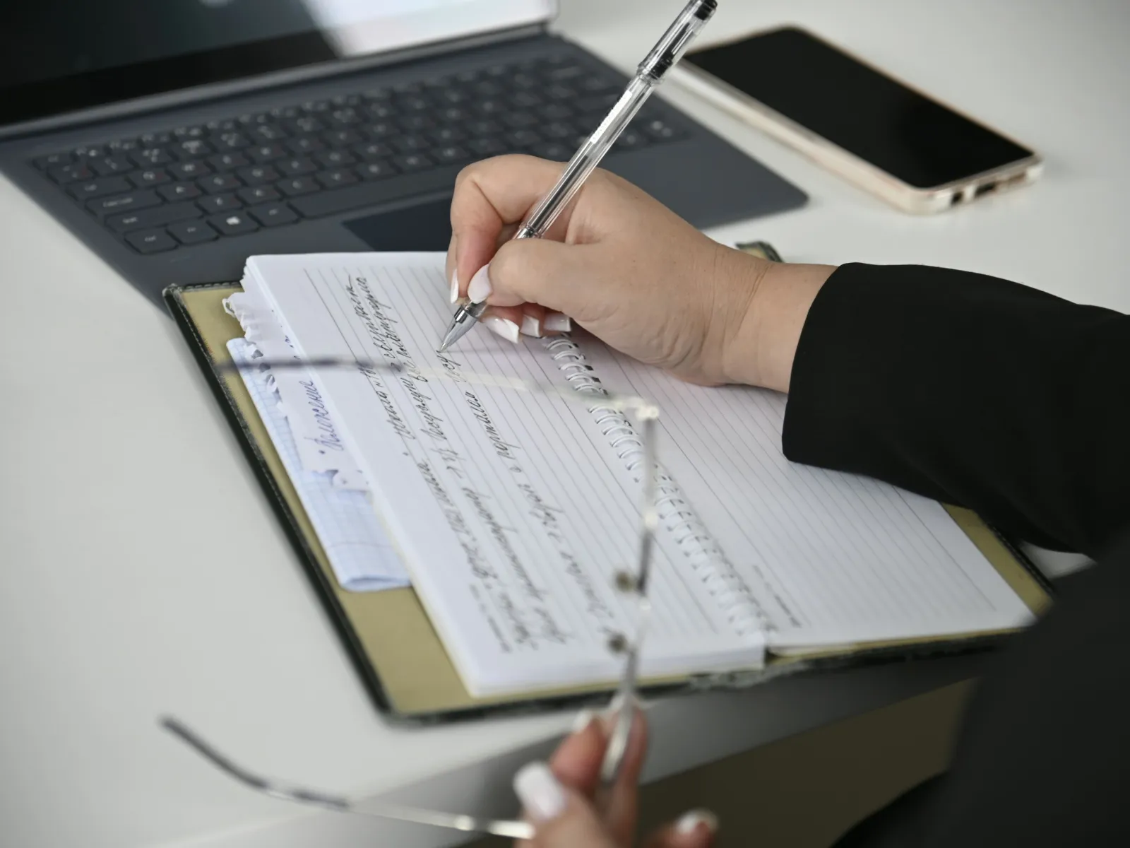Person writing notes in a lined notebook with a laptop and smartphone on a white desk.