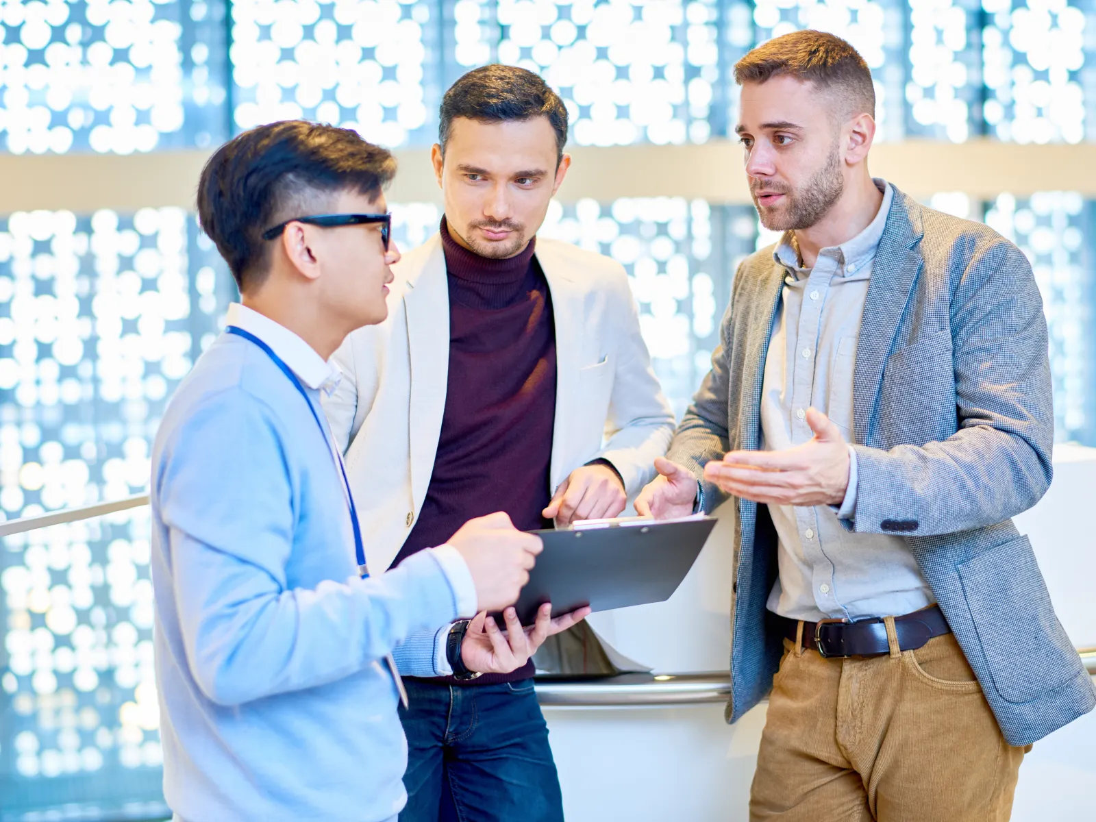Three young professionals engaged in discussion with clipboard in a modern office setting with patterned windows.