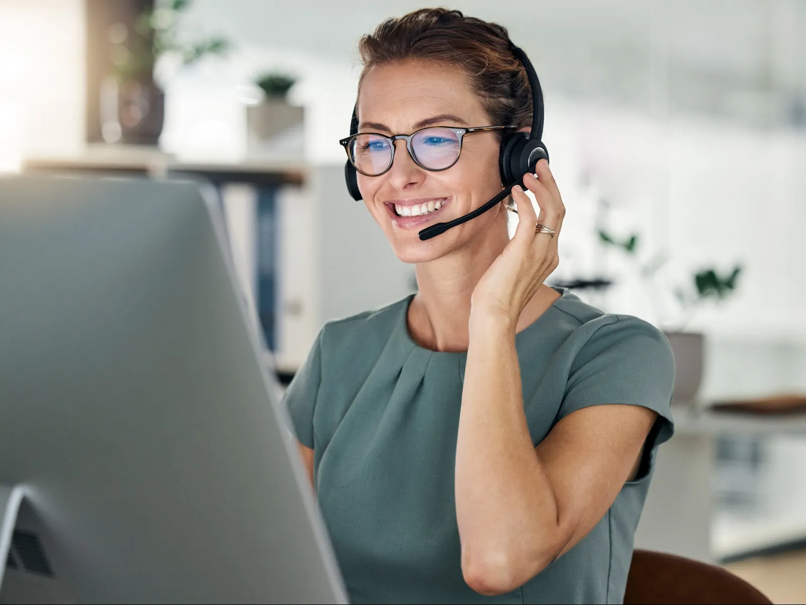 Smiling woman wearing headset and glasses working on a desktop computer in bright modern office