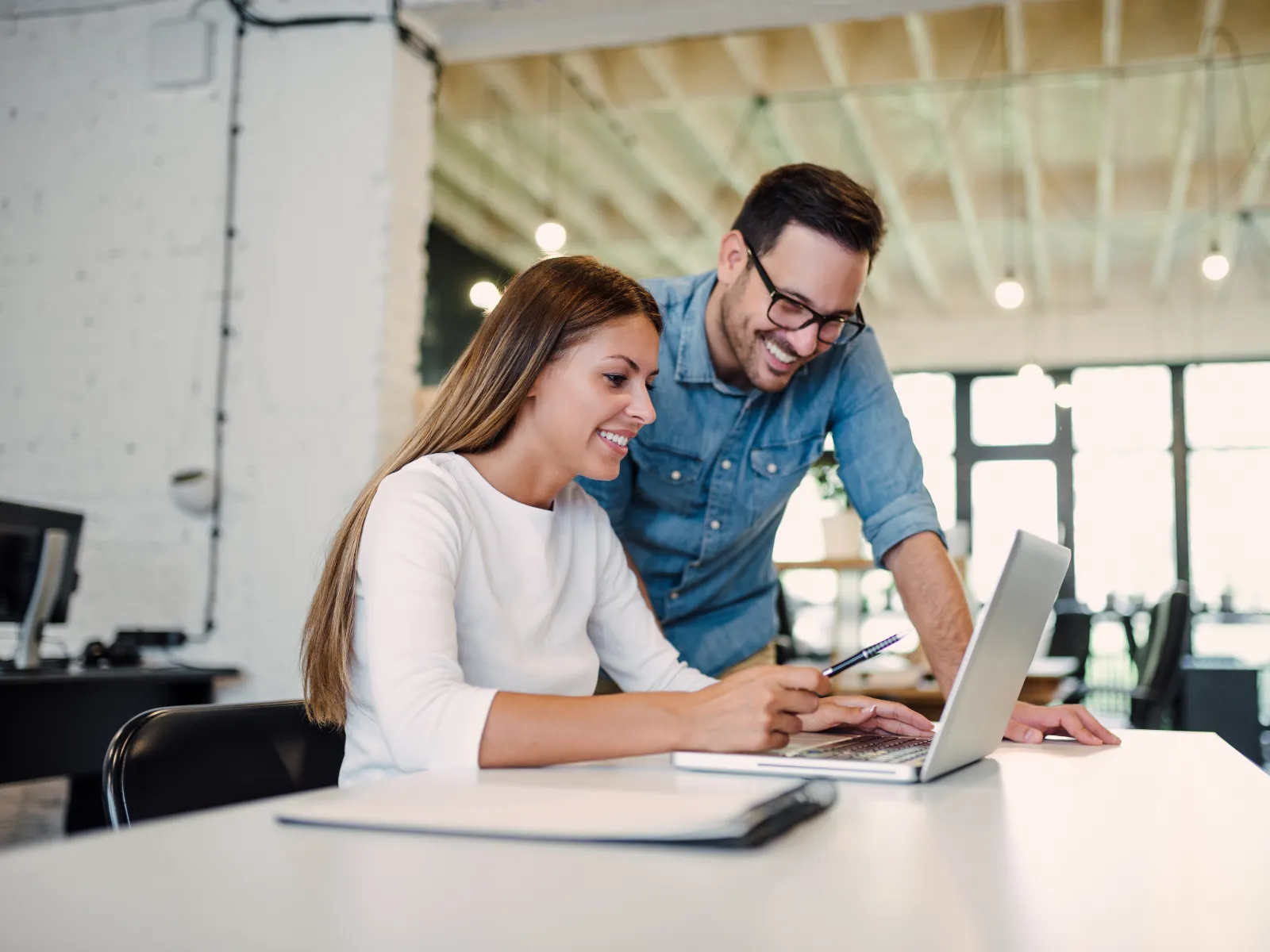 Two colleagues smiling and collaborating on a laptop in a modern bright office space.